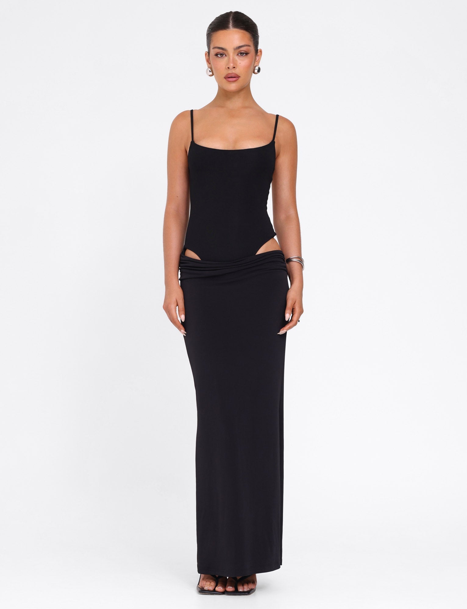 A woman faces forward in the TIGERMIST UMA DRESS - BLACK, a sleeveless fitted maxi with thin adjustable straps and hip cutouts. She has her hair pulled back, wears hoop earrings, bracelets, black sandals, and stands against a plain white background.