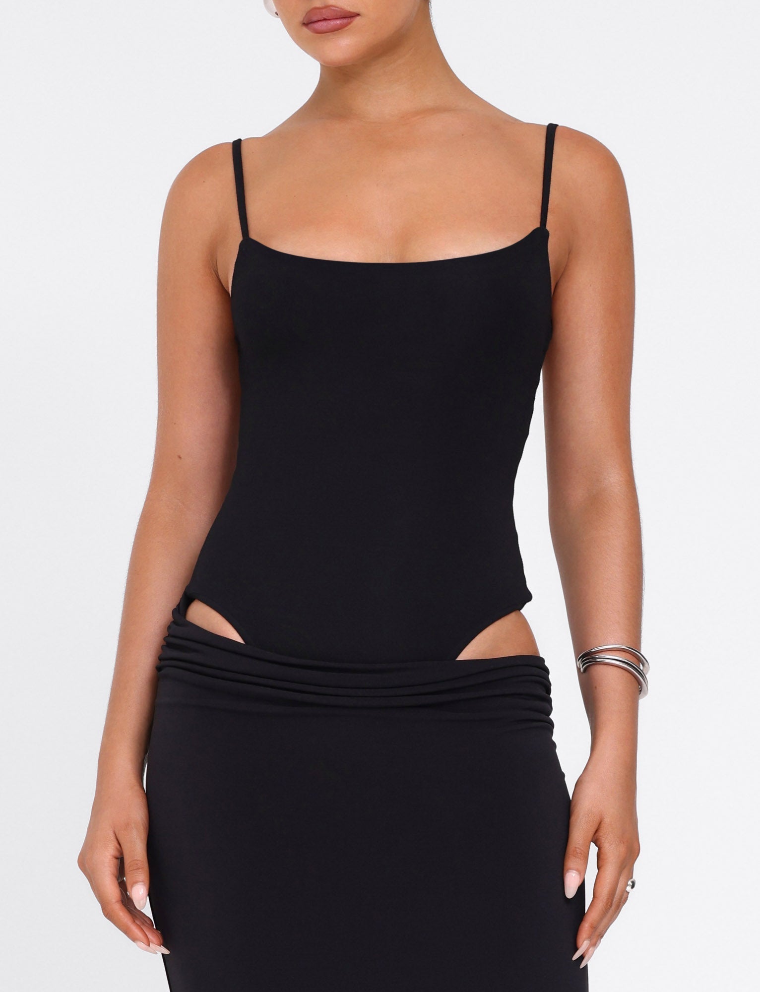 A woman wears the UMA DRESS - BLACK by TIGERMIST, a bodycon maxi dress with adjustable shoulder straps, paired with a black skirt with side waist cutouts. She accessorizes with silver bangles on her left wrist and has a neutral expression.