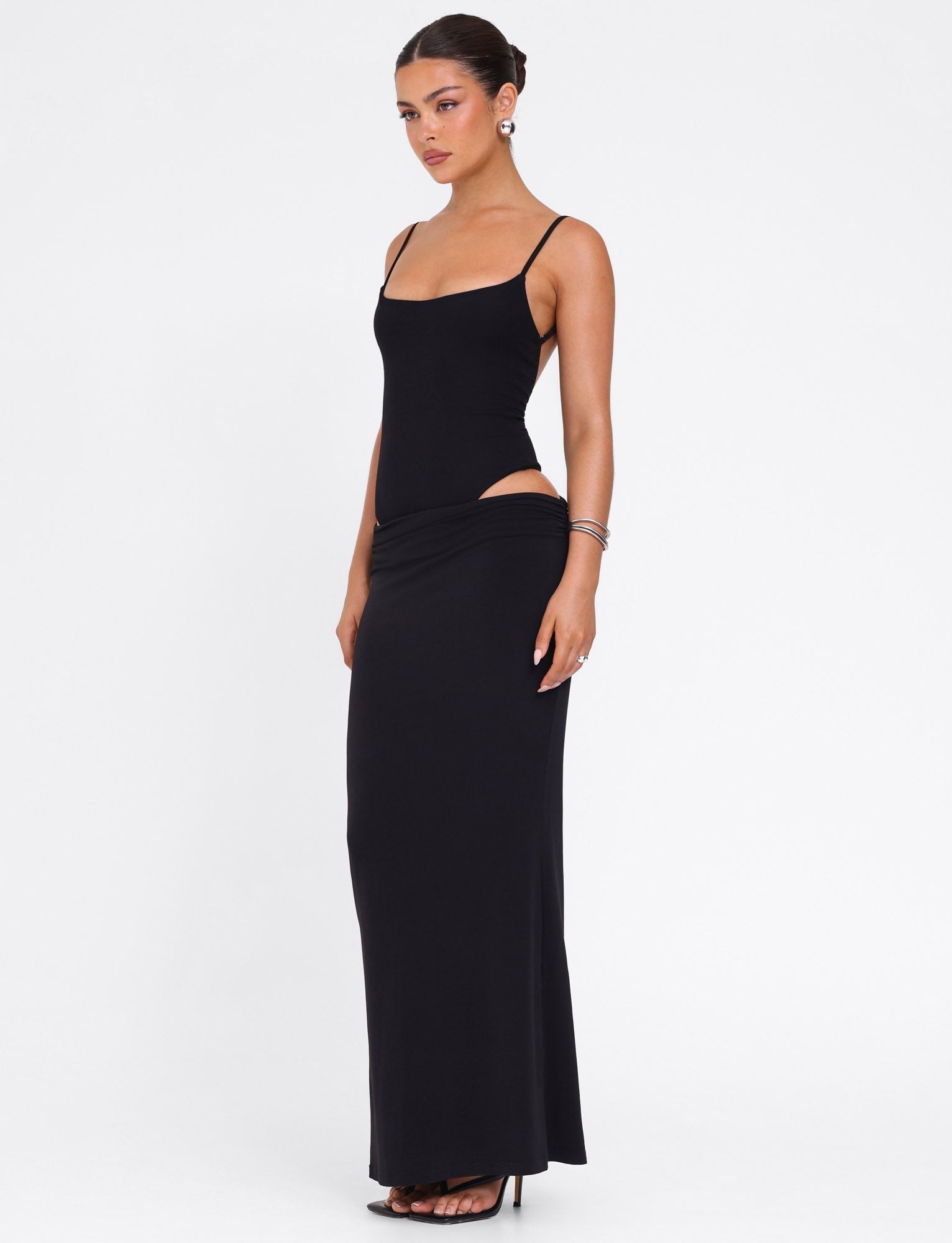 A woman wears the TIGERMIST UMA DRESS - BLACK, a sleek black bodycon maxi with adjustable straps and high side cutouts, made from high stretch jersey. She pairs it with earrings, bracelets, heels, and has her hair pulled back against a white backdrop.