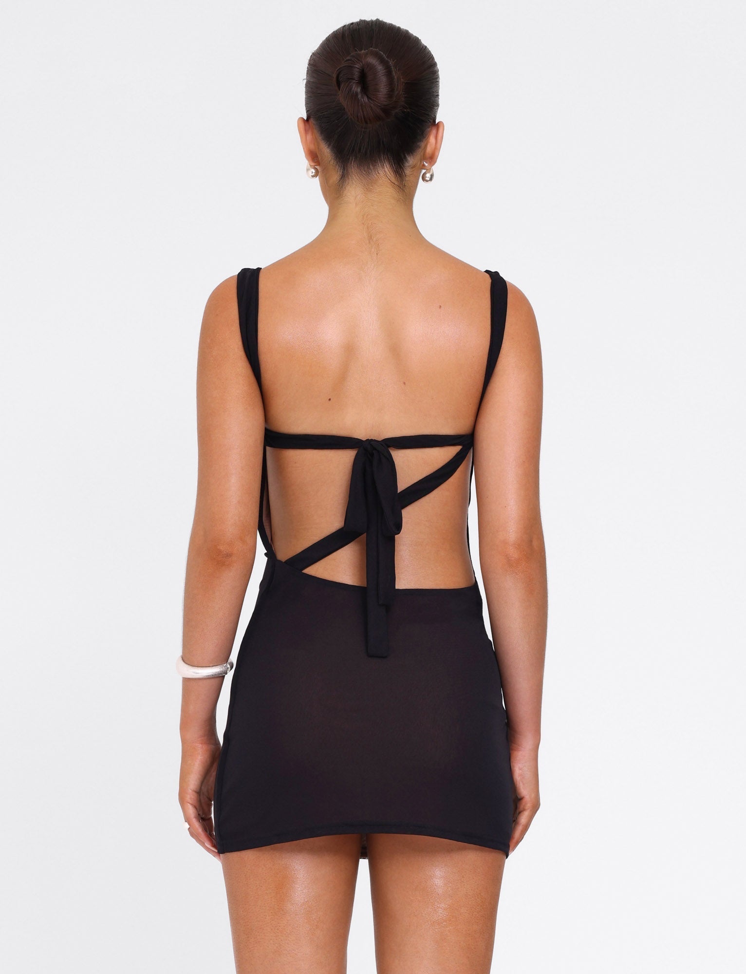 A woman with her hair in a bun wears the TIGERMIST FREYA DRESS - BLACK, a mini dress with an open back and tie detail made from buttery soft fabric, standing against a plain white background.