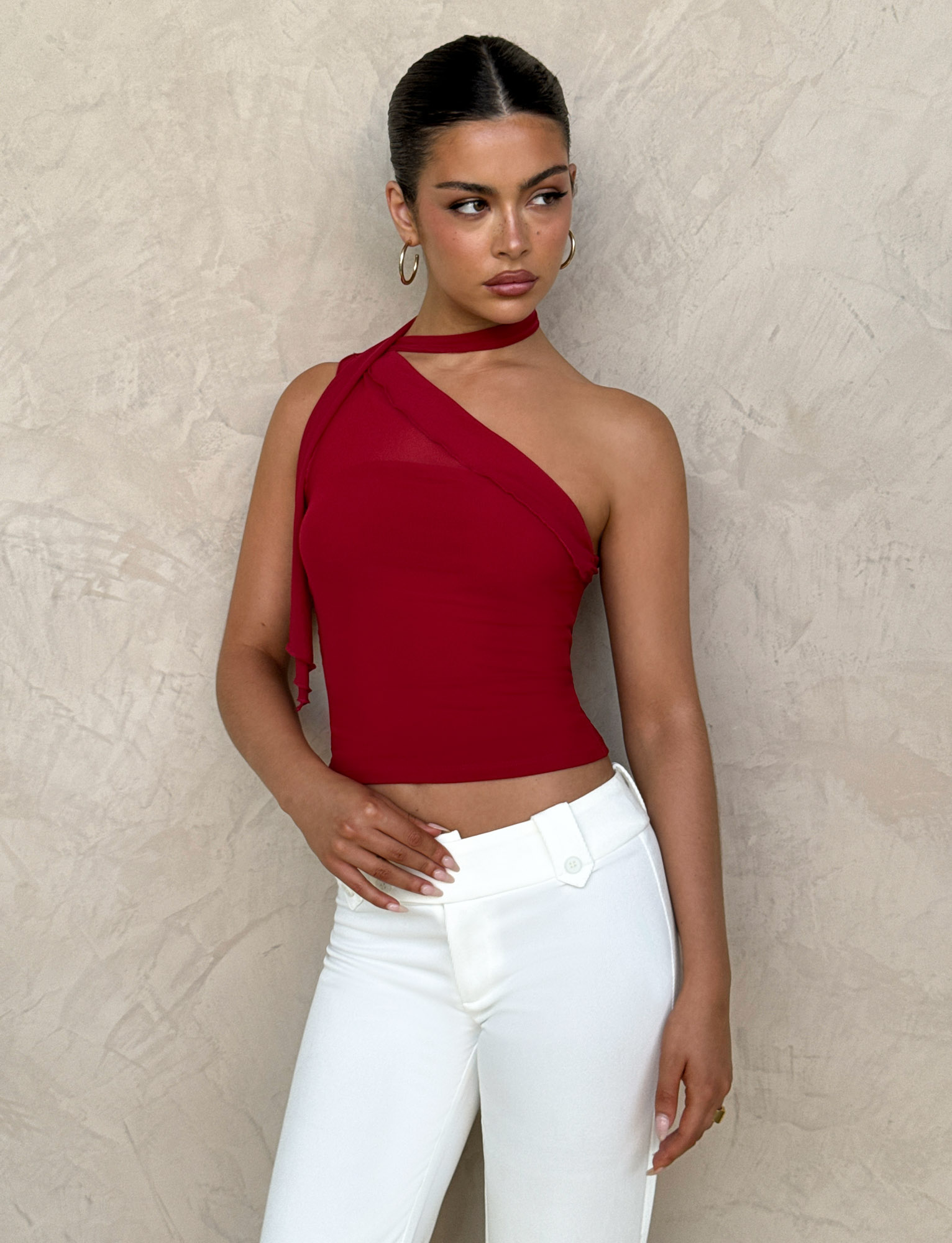 A woman wears the TIGERMIST FIONA TOP in red with white pants, standing against a textured beige wall, looking to her left with one hand on her hip.