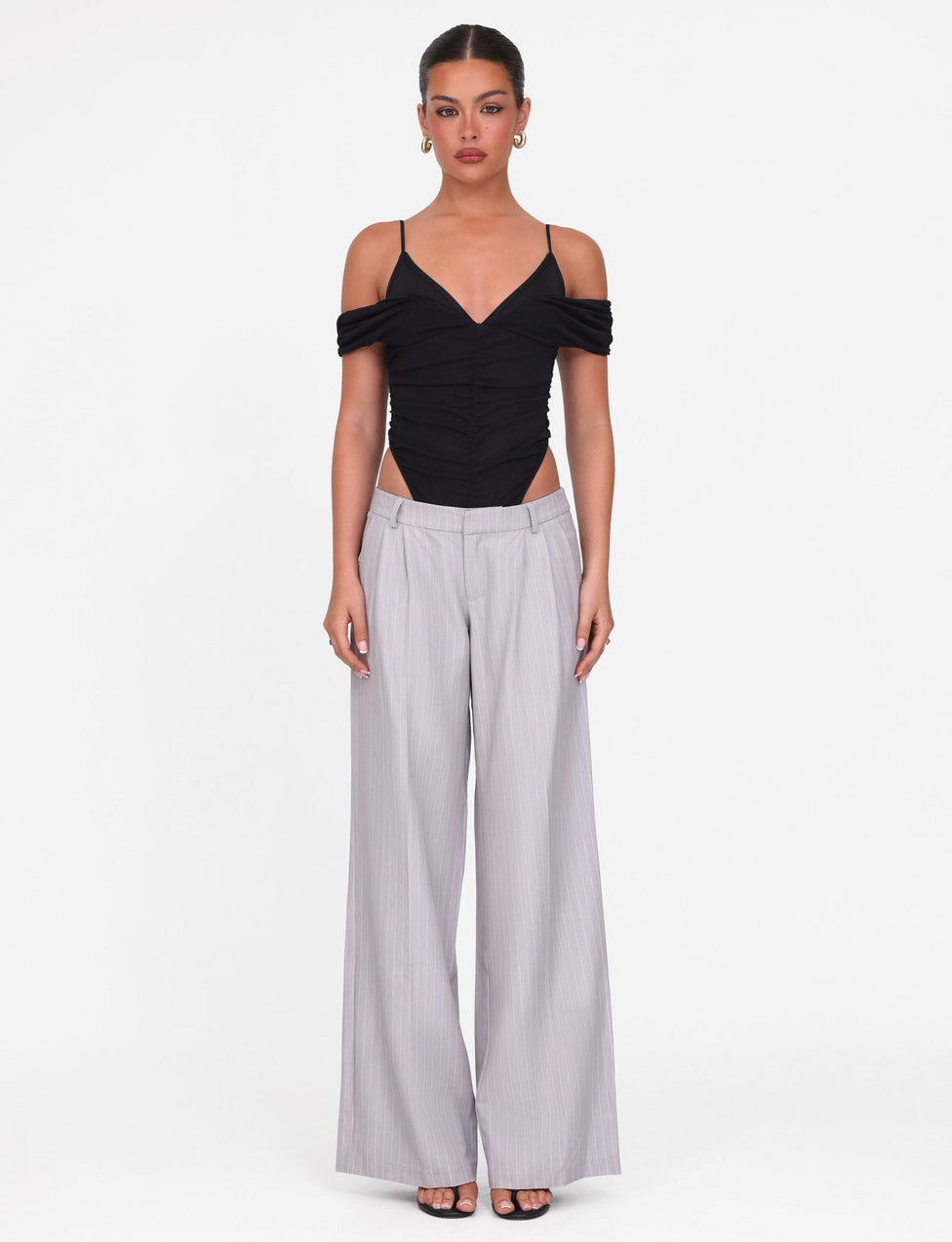 A woman faces forward with a neutral expression, wearing a black off-shoulder spaghetti strap top and TIGERMIST’s AMBERLY SUIT PANT in grey pinstripe against a white background.