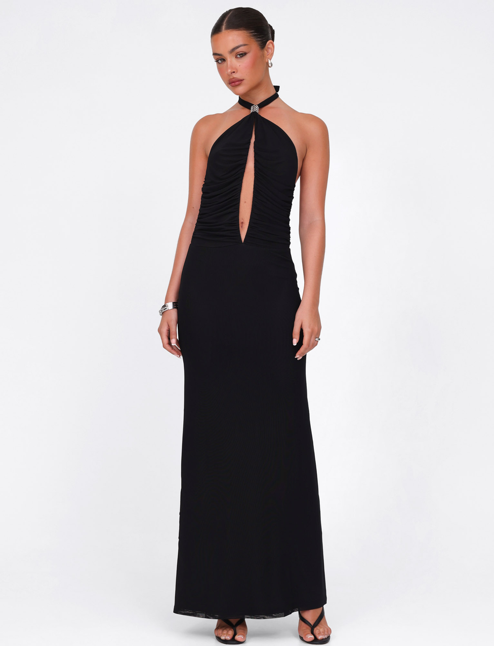 A woman wears the TIGERMIST ZARA MAXI DRESS in black, featuring a front keyhole cutout. She stands against a plain white background with her hair pulled back, accessorized with bracelets and earrings.