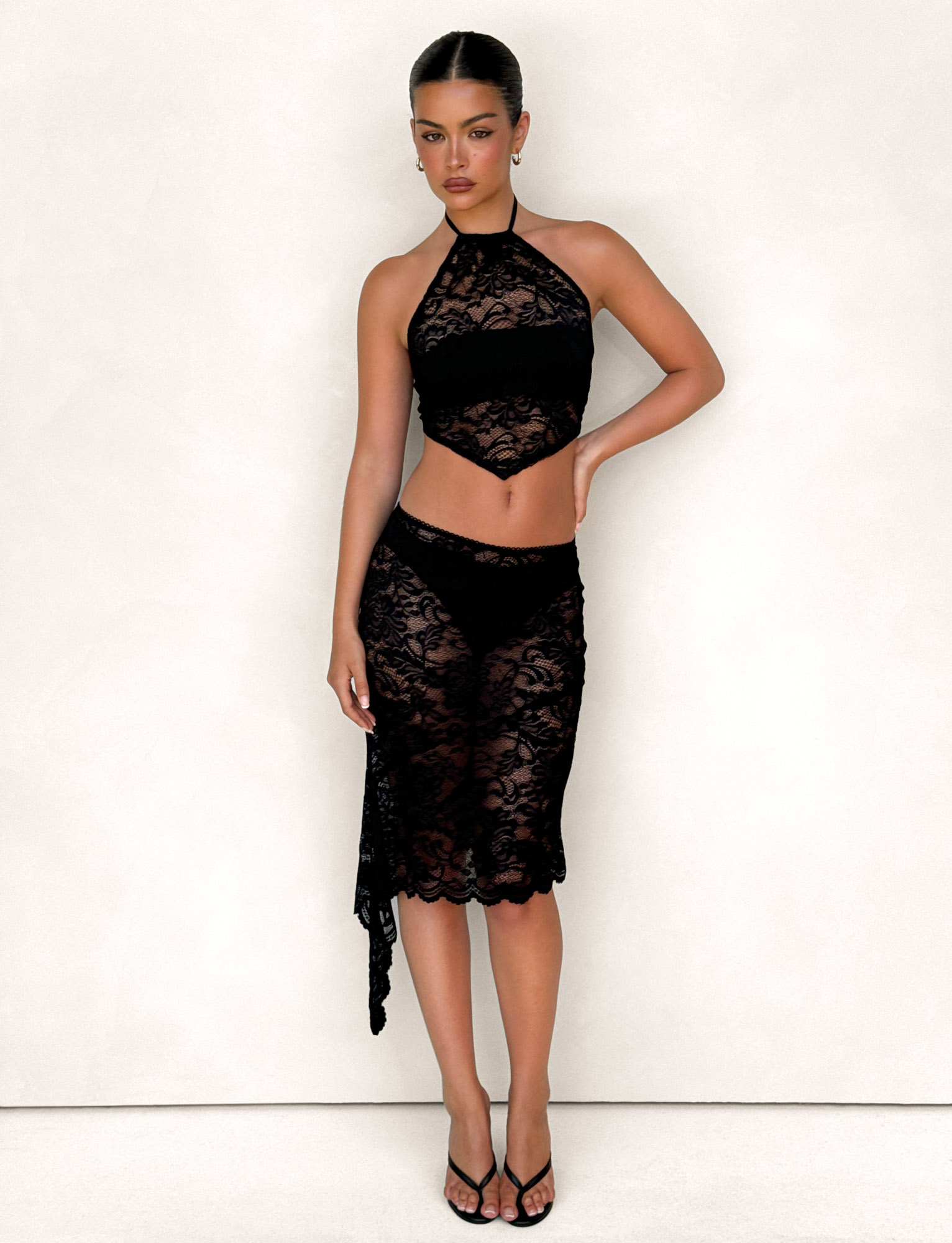 Against a light beige background, a woman with sleek, pulled-back dark hair wears the TIGERMIST SIYA MIDI SKIRT in black lace and a matching black halter crop top, styled with black high-heeled sandals.
