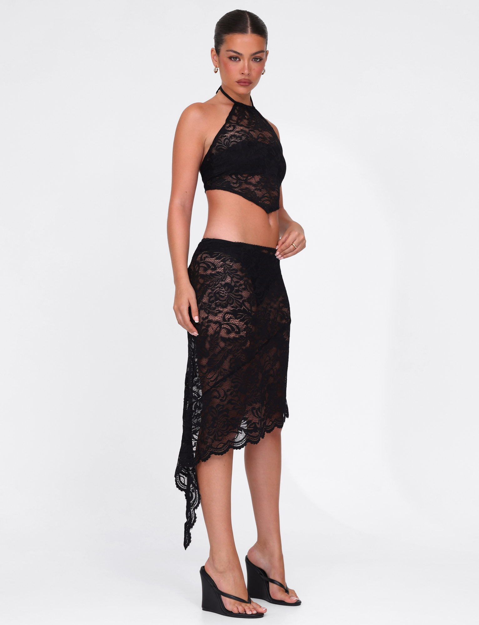 A woman models the TIGERMIST SIYA MIDI SKIRT in black lace, featuring a sheer finish and asymmetrical hemline, paired with a matching halter top and black wedge sandals against a plain white backdrop.