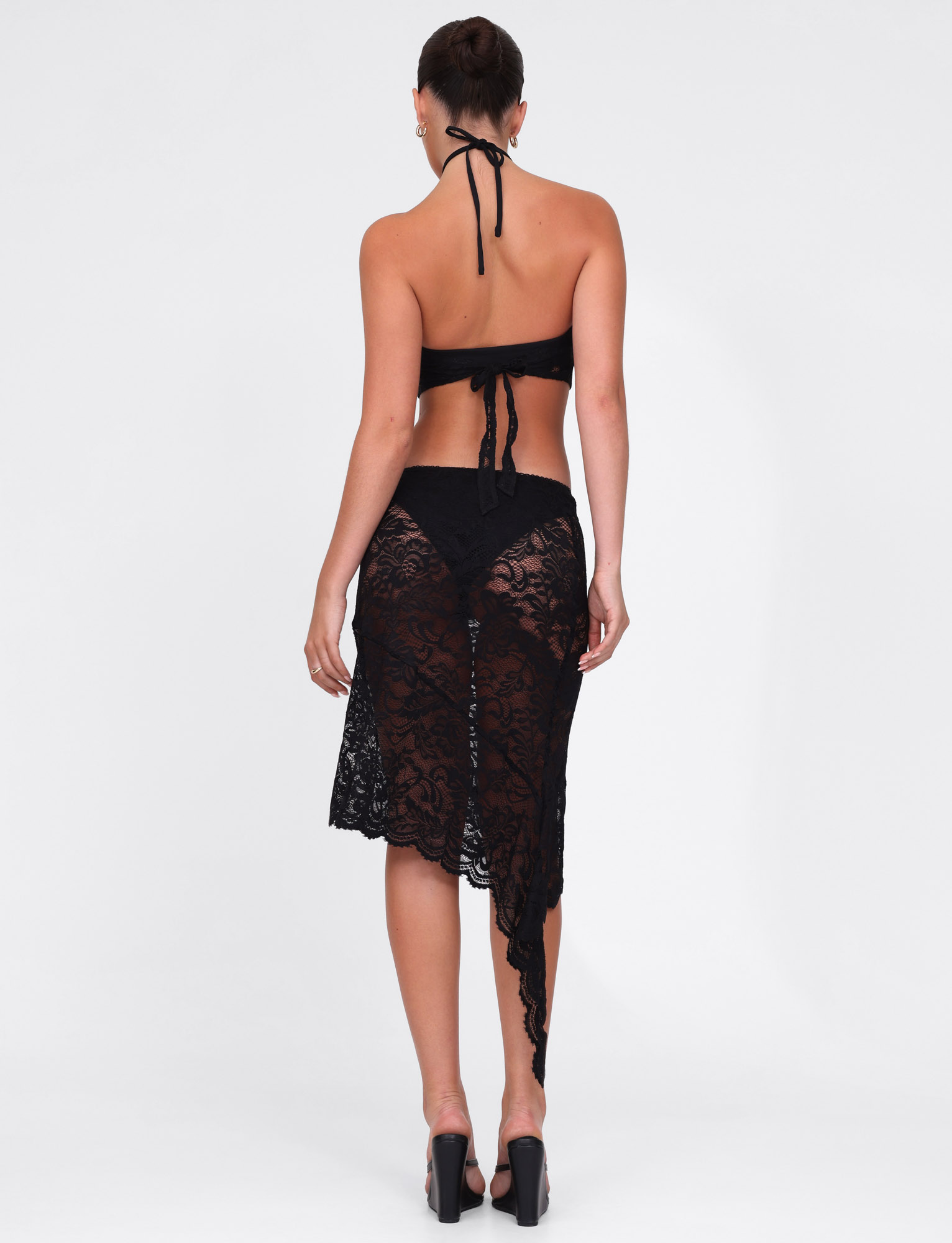 A woman faces away wearing the TIGERMIST SIYA MIDI SKIRT in black lace over black underwear, paired with a black halter crop top and black high heels, set against a plain white background.