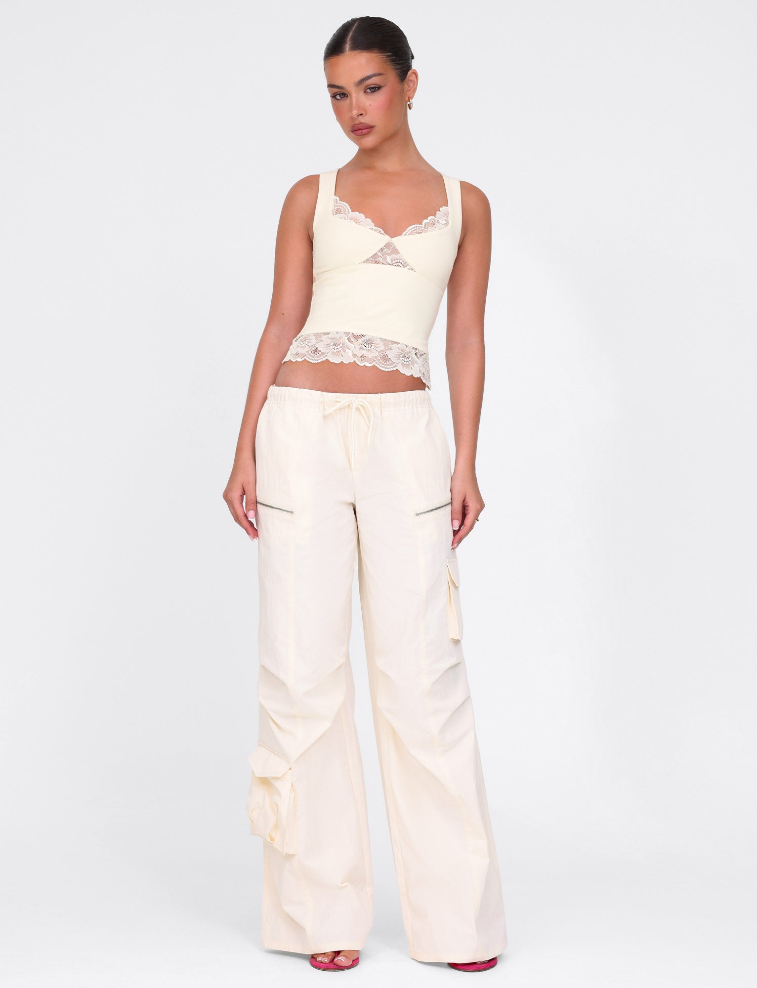A person in a DIONE LACE TOP - YELLOW : BUTTER : SOFT BUTTER by TIGERMIST, paired with wide-legged drawstring pants and red open-toed shoes, stands against a white background with their hands by their sides.