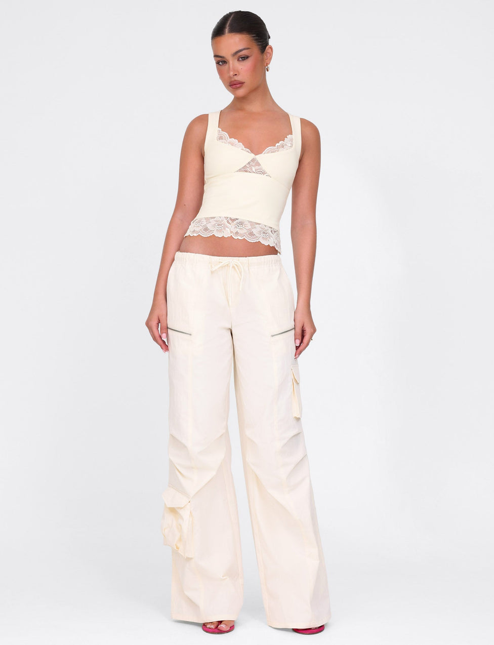 A person in a DIONE LACE TOP - YELLOW : BUTTER : SOFT BUTTER by TIGERMIST, paired with wide-legged drawstring pants and red open-toed shoes, stands against a white background with their hands by their sides.