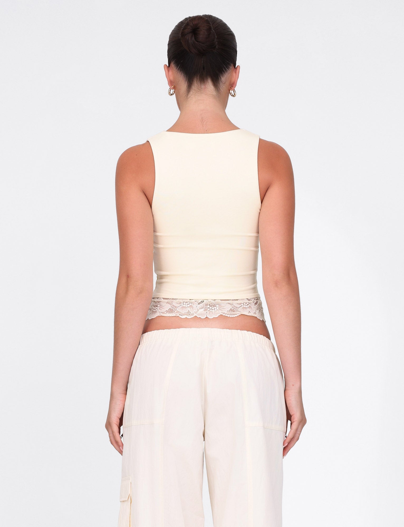 A person with dark hair styled in a bun is seen from behind wearing TIGERMIST's DIONE LACE TOP in soft butter yellow. The sleeveless jersey top is adorned with delicate lace trim at the hem. Paired with light pants, the minimalist ensemble stands out against a crisp white backdrop.