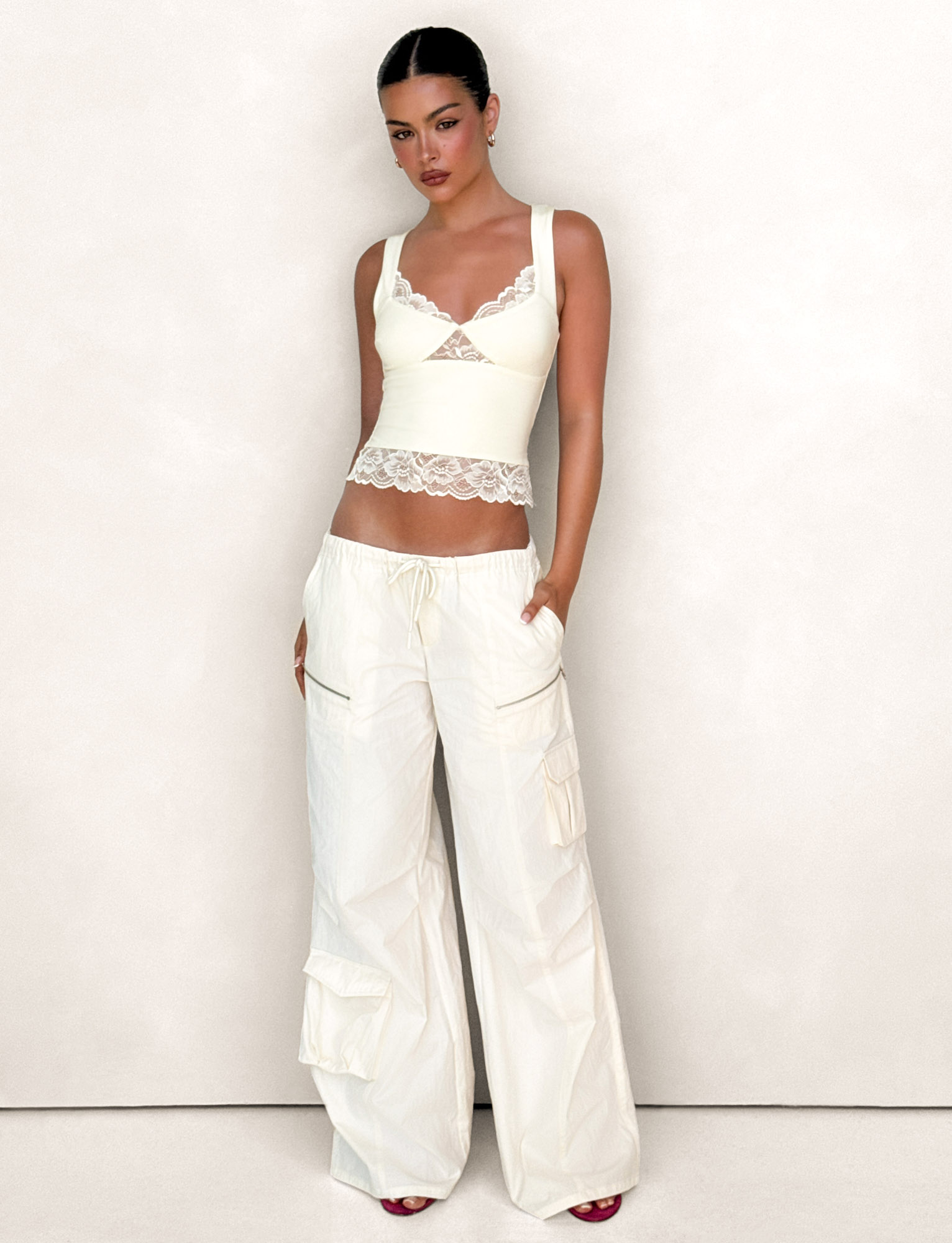 A woman with dark hair pulled back and dark lipstick poses against a light background, wearing TIGERMIST's HERMIONE PANT in Yellow Butter with functional cargo pockets and a cream lace-trim tank top.