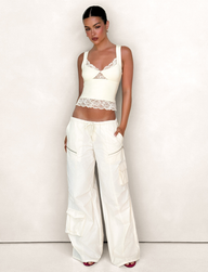 A woman with dark hair pulled back and dark lipstick poses against a light background, wearing TIGERMIST's HERMIONE PANT in Yellow Butter with functional cargo pockets and a cream lace-trim tank top.