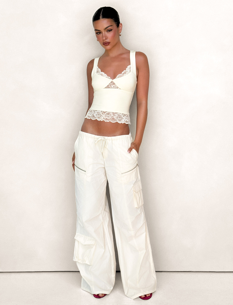 A woman with dark hair pulled back and dark lipstick poses against a light background, wearing TIGERMIST's HERMIONE PANT in Yellow Butter with functional cargo pockets and a cream lace-trim tank top.