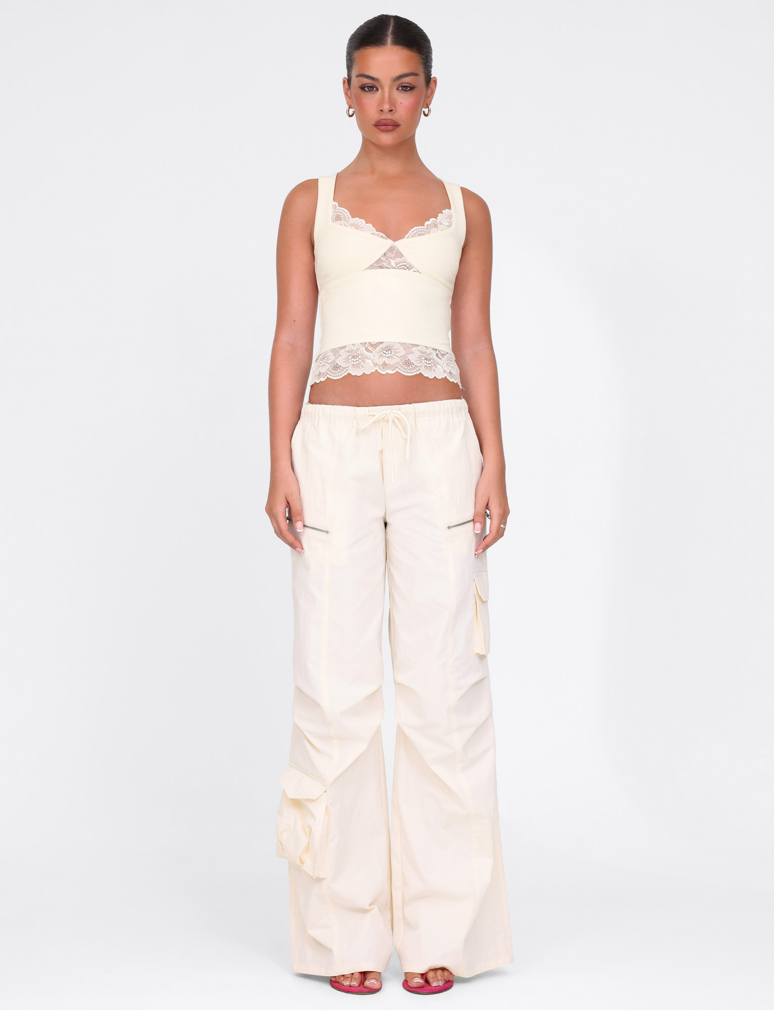 A woman stands against a white background wearing TIGERMIST's HERMIONE PANT in Soft Butter, featuring a relaxed fit and functional pockets. She pairs the yellow cargo pants with a cream crop top and red heels, her hair pulled back, gazing neutrally ahead.