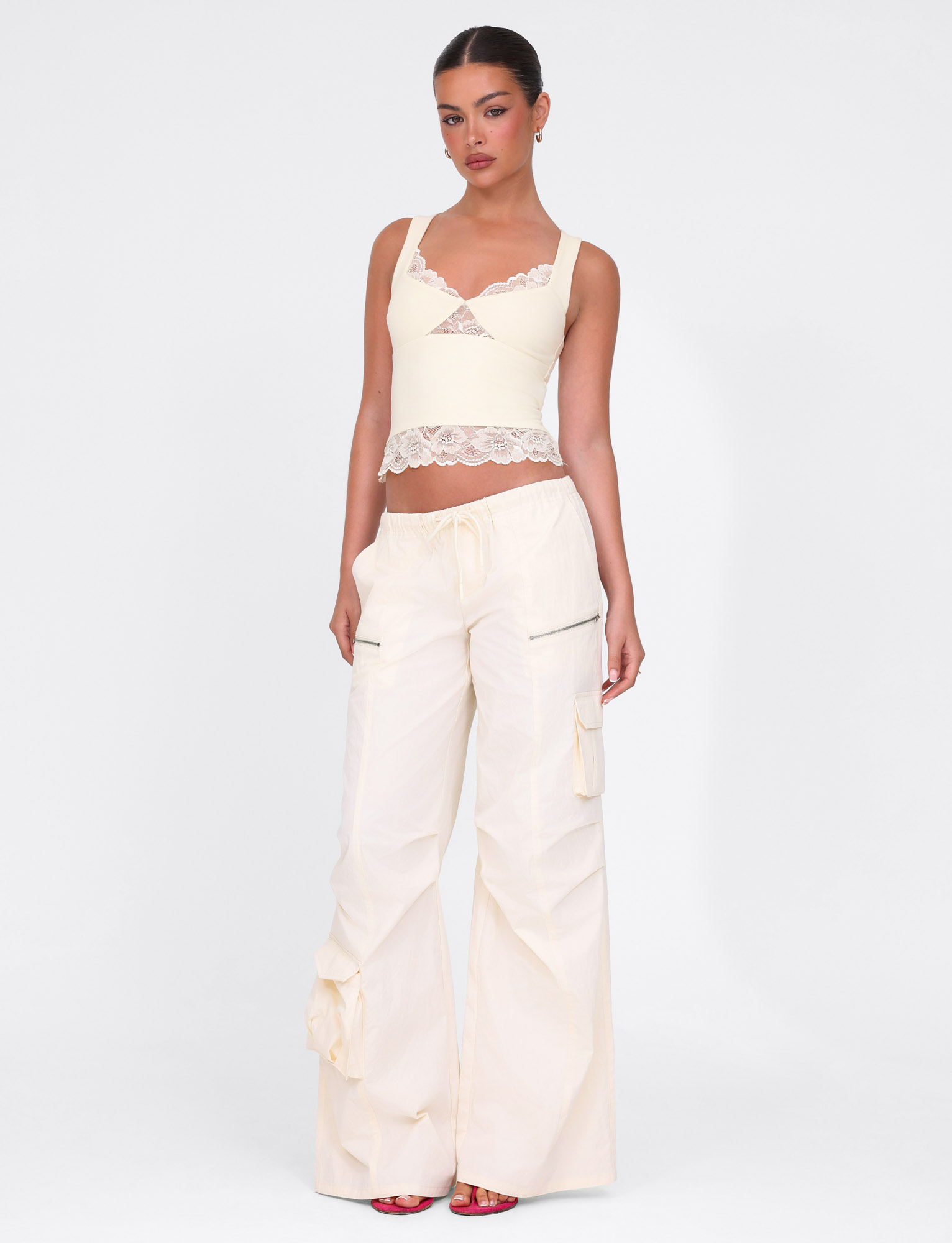 A woman stands against a white background in TIGERMIST HERMIONE PANT - YELLOW: BUTTER : SOFT BUTTER, featuring an elasticated waist, side zippers, and pockets. She pairs them with a cream lace-trim tank top and red open-toe heels.
