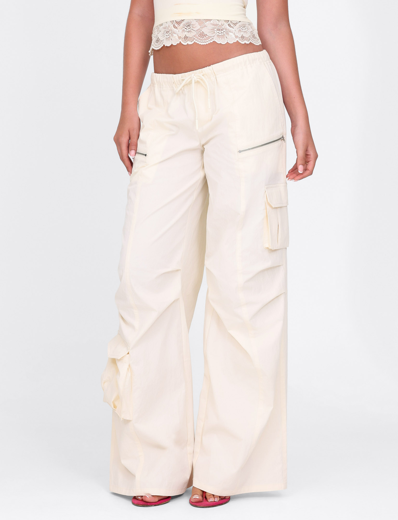 A person wears TIGERMIST's HERMIONE PANT in Soft Butter, featuring a yellow, cream-toned color, cargo pockets, and an elasticated waist, paired with a lace-trimmed cropped top and red heeled sandals.