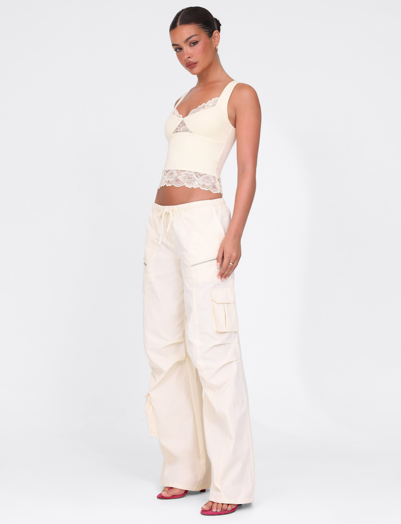 A woman models the TIGERMIST HERMIONE PANT in soft butter yellow, paired with a lace-trimmed cream crop top and pink heeled sandals, standing against a plain white background.