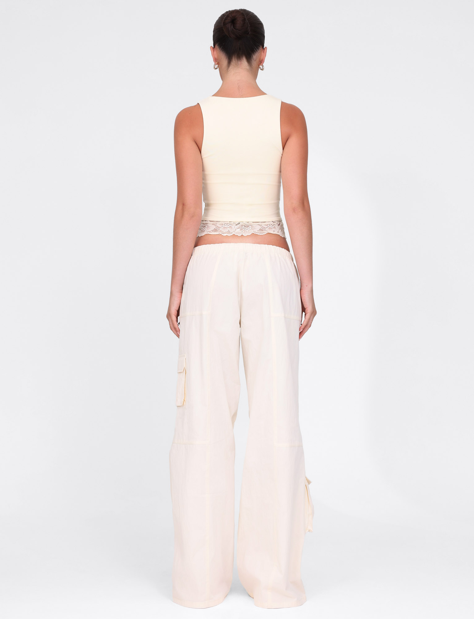 A person stands facing away from the camera, wearing TIGERMIST's HERMIONE PANT in Soft Butter Yellow, featuring lace details at the elasticated waist, paired with a sleeveless cream tank top against a plain white background.