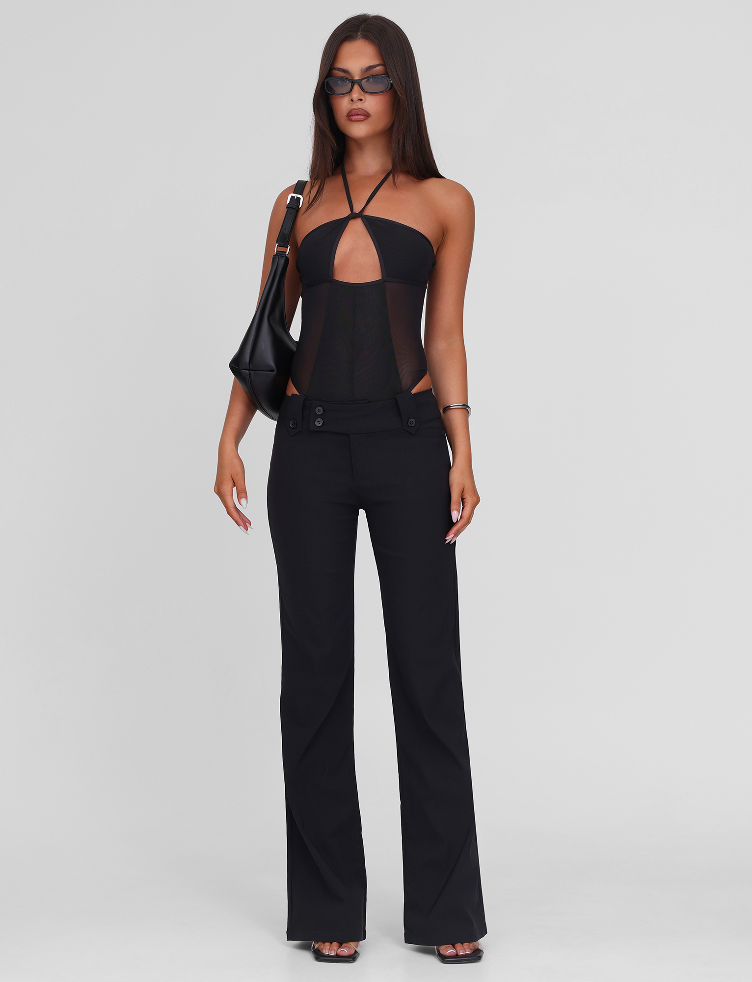 A woman poses against a white background wearing the TIGERMIST MELANIA BODYSUIT - BLACK, paired with high-waisted stretch black pants, heeled sandals, dark sunglasses, and a black shoulder bag.