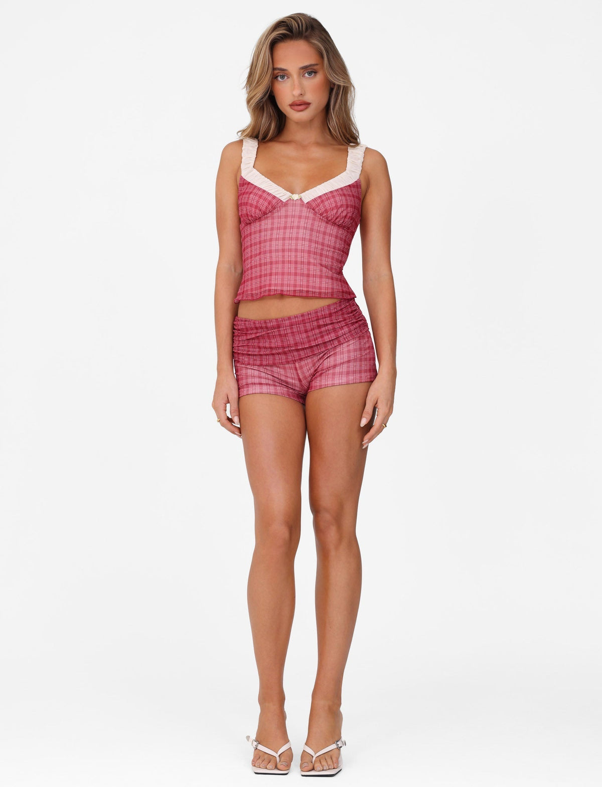Against a white background, a woman sports the TIGERMIST AMARAN SHORT in RED CHECK, featuring a sleeveless top with lace trim and matching shorts. Her chic look is completed with blonde hair and white heeled sandals.