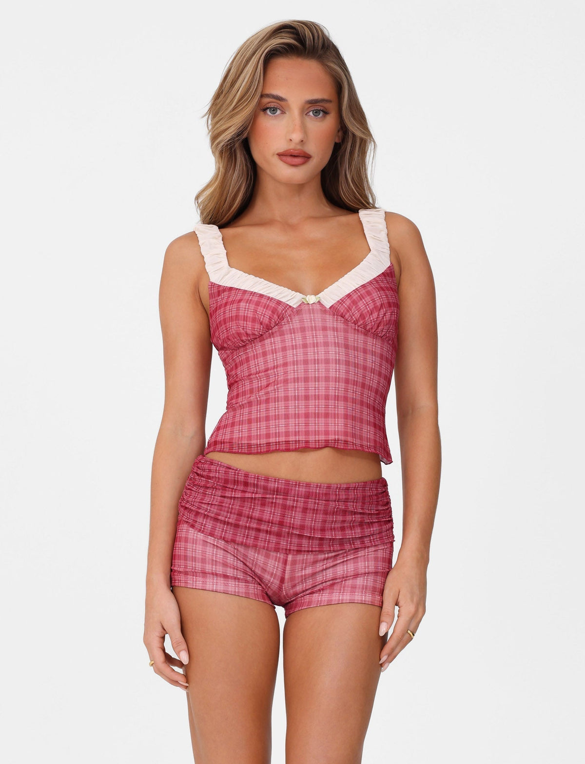 A person models the Tigermist AMARAN SHORT - RED: RED CHECK against a white background, featuring a red check print crop top with white ruffled straps and matching high-waisted shorts. Their long, wavy hair complements the bodycon fit as they maintain a neutral expression.