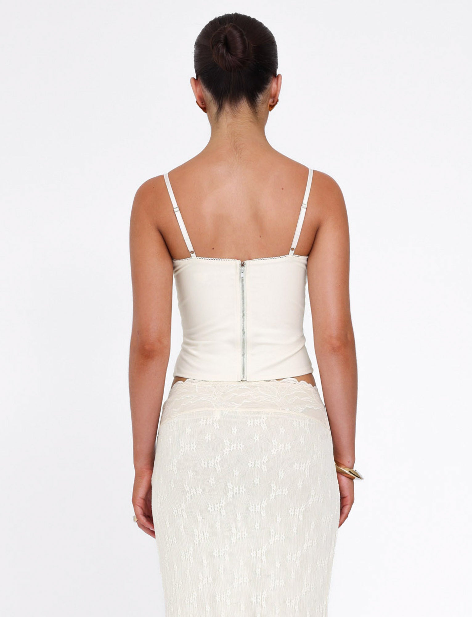 A woman with her hair in a neat bun faces away from the camera, wearing the TIGERMIST ANTONELLA TOP in white, bone, or cream and a sheer lace panel skirt. The background is plain white.