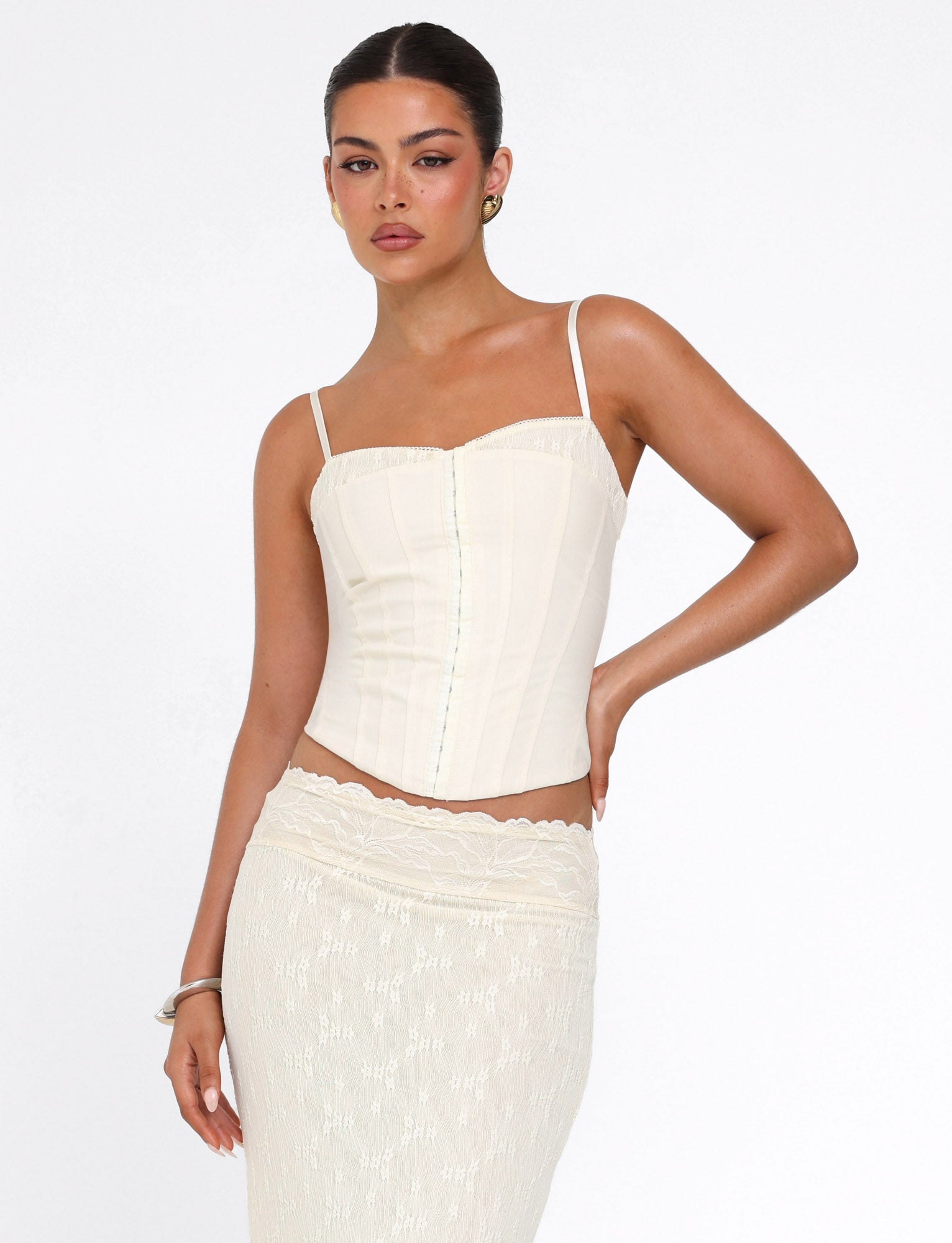 A woman poses against a white background wearing the TIGERMIST ANTONELLA TOP in bone cream, a fitted suiting corset with thin straps, paired with a matching lace-hem skirt and one hand on her hip.