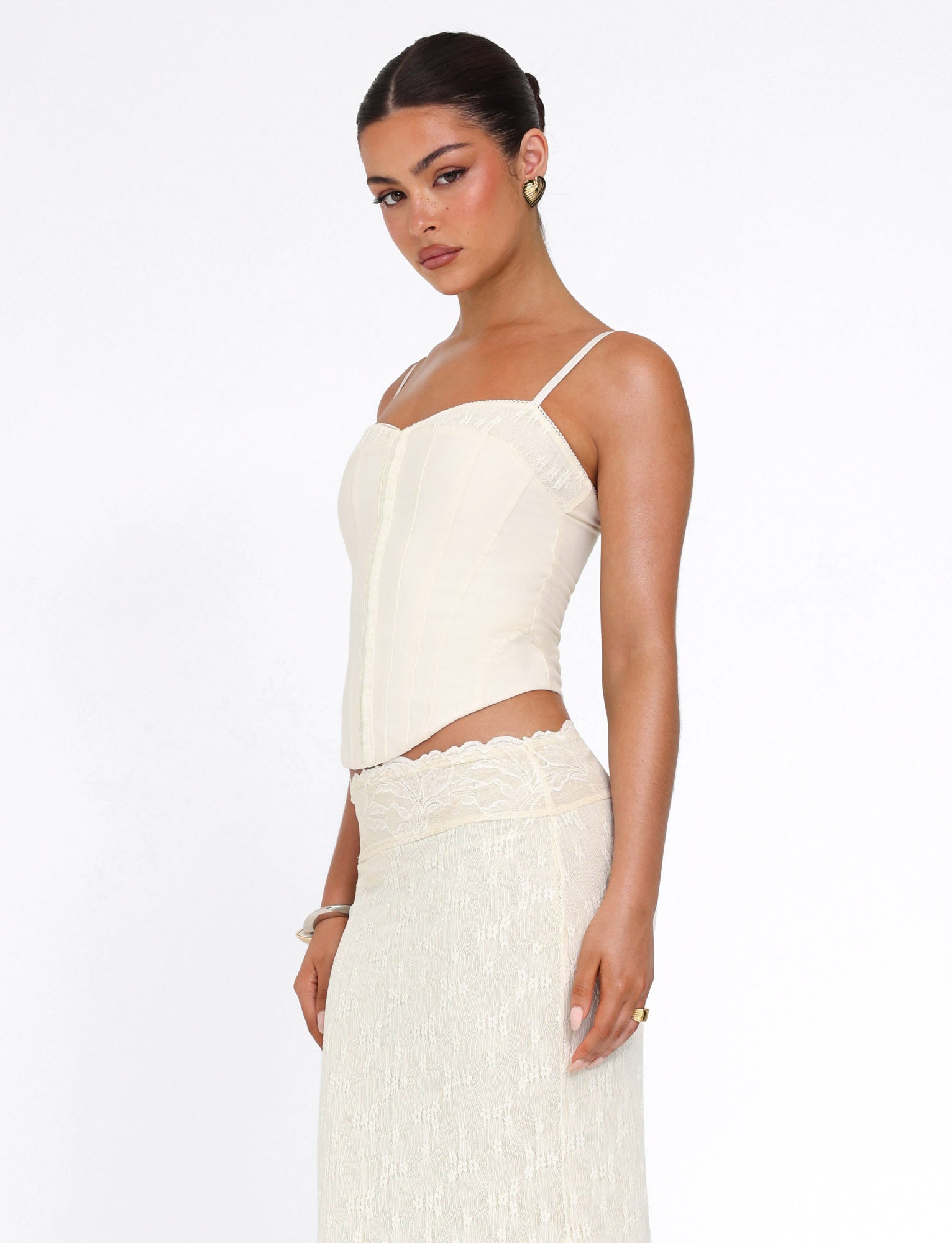 A woman wears the TIGERMIST ANTONELLA TOP in cream, featuring delicate lace panels, paired with a matching lace skirt. She stands against a plain white background, looking slightly to the side with a neutral expression.