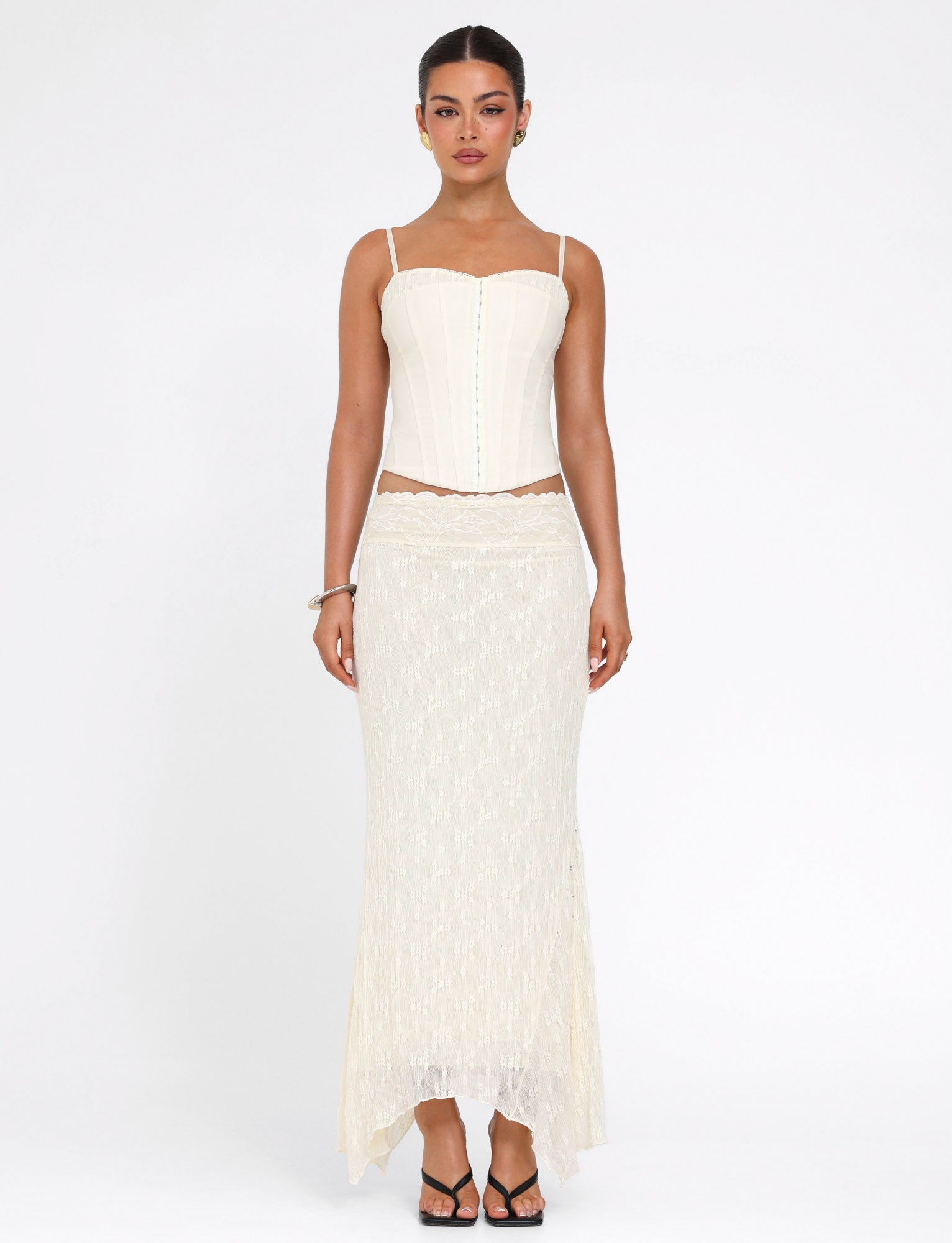 A woman stands against a white backdrop wearing the TIGERMIST ANTONELLA MIDI SKIRT in cream lace and a matching cream corset top. Her hair is pulled back and she accessorizes simply with a bracelet.