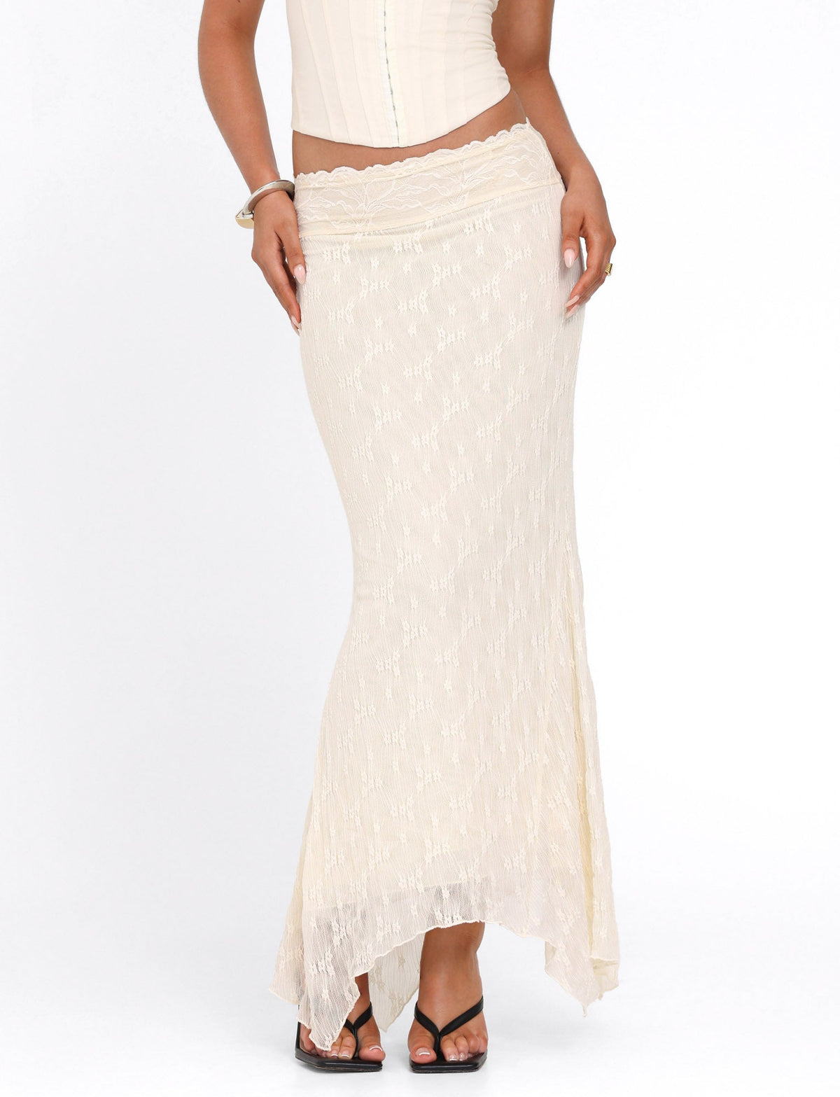 A woman wears the TIGERMIST ANTONELLA MIDI SKIRT in cream lace and an asymmetric hem, paired with a matching lace corset and black strappy heels, standing against a white background.