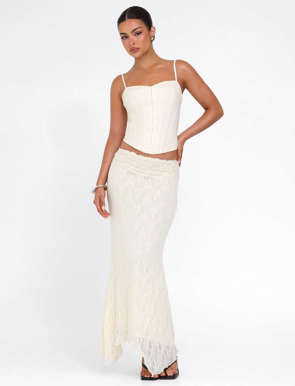 A woman poses confidently against a white background wearing the TIGERMIST ANTONELLA MIDI SKIRT in cream lace, paired with a matching cream corset top, accessorized with earrings, a bracelet, and black sandals.