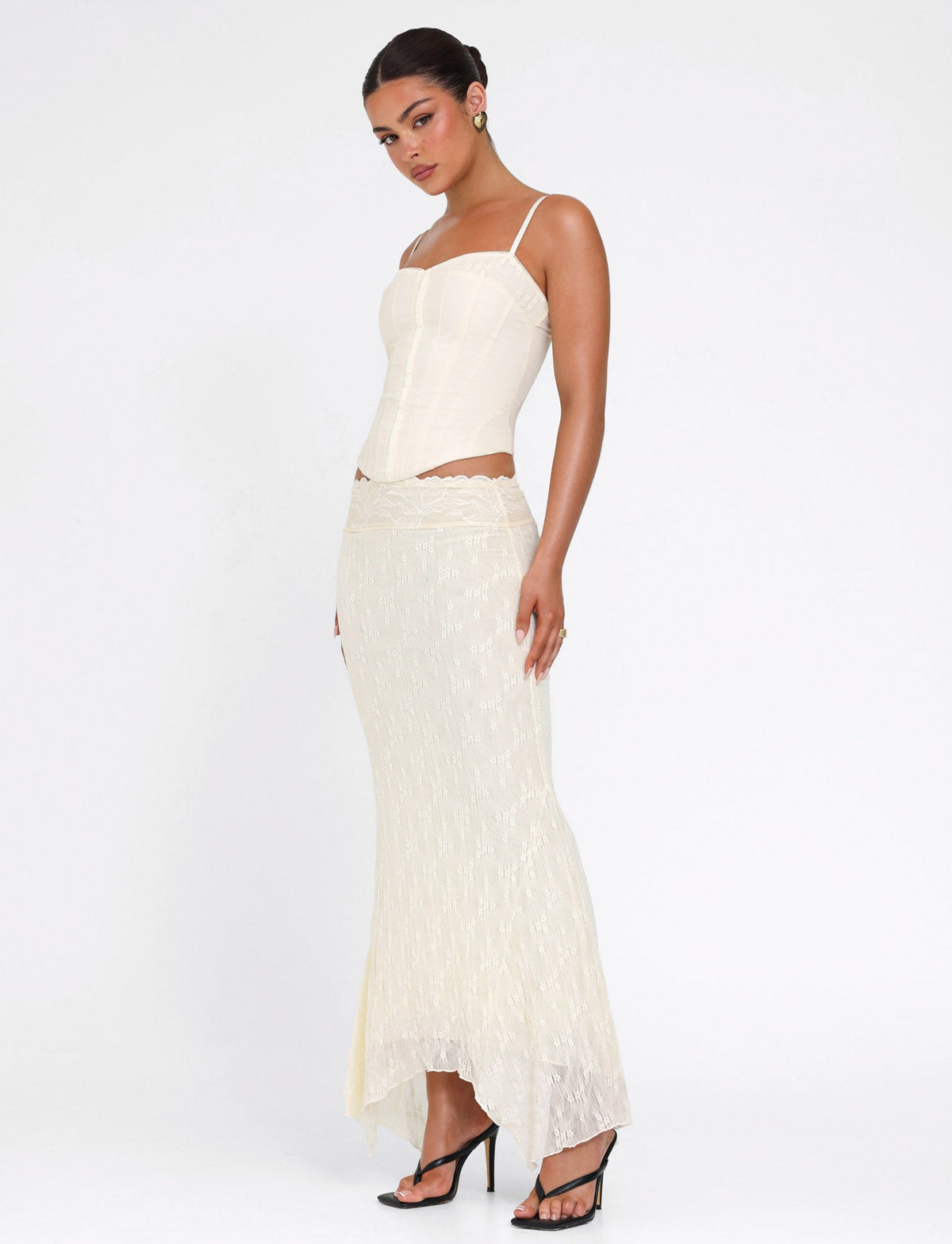A woman stands against a white background wearing the TIGERMIST ANTONELLA MIDI SKIRT in cream lace, styled with a matching corset top, black heeled sandals, and her hair pulled back.