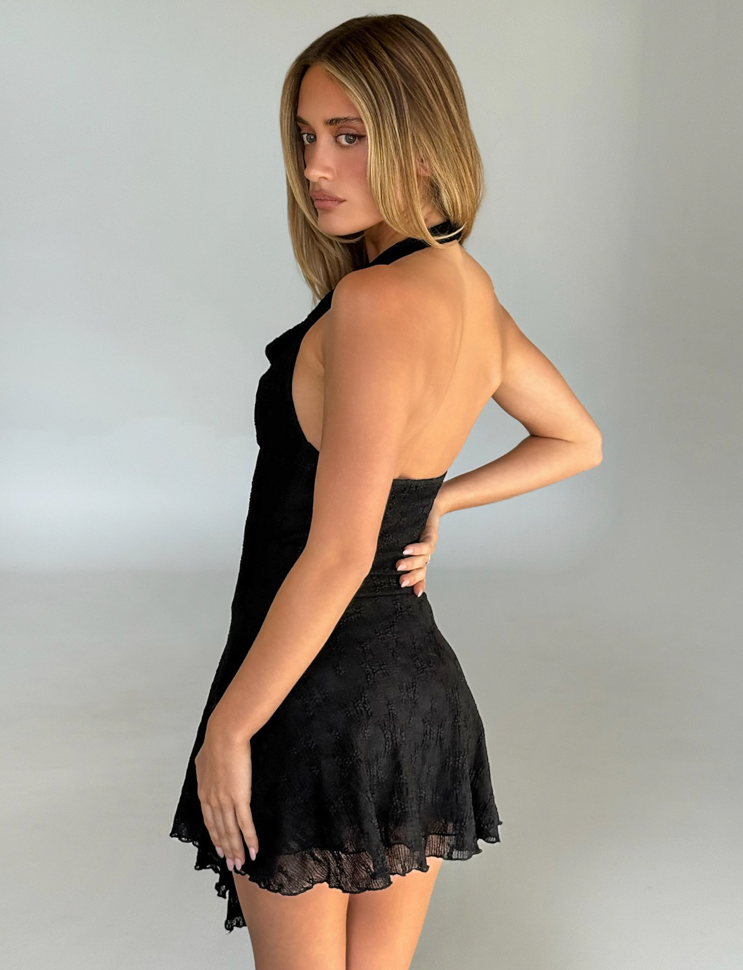 A woman with straight, light brown hair looks over her shoulder at the camera, wearing the TIGERMIST FIORELLA DRESS in black lace. The sleek mini dress features a halter neckline and subtle texture against a plain, light background.