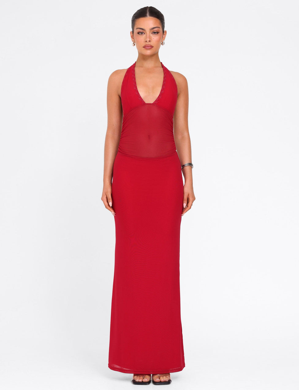 A woman faces forward in the TIGERMIST BRESILY MAXI DRESS - RED, a sleeveless, floor-length gown with a sheer midsection and elegant embroidery. She wears earrings and a bracelet, hair pulled back, set against a plain white background.