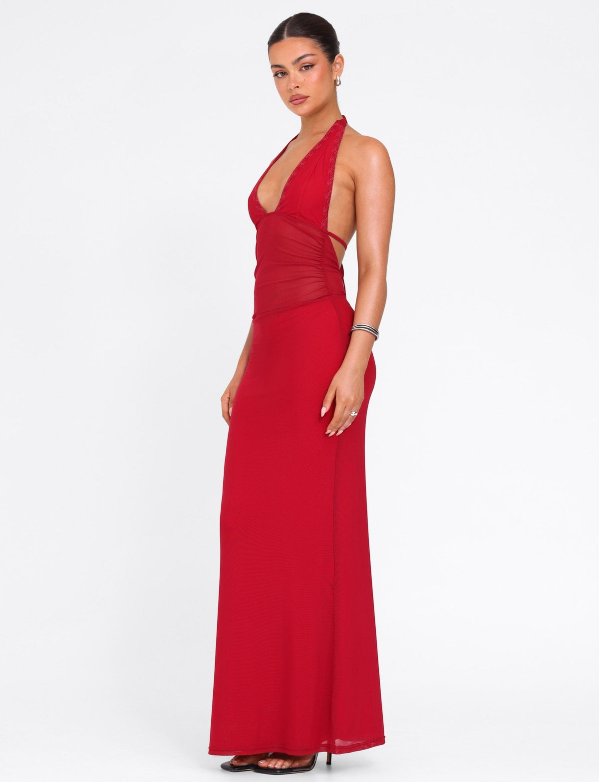 A woman stands against a white background wearing the TIGERMIST BRESILY MAXI DRESS in red, featuring tie halter straps and a fitted bodice. Her hair is tied back, paired with earrings, a bracelet, and black heels.