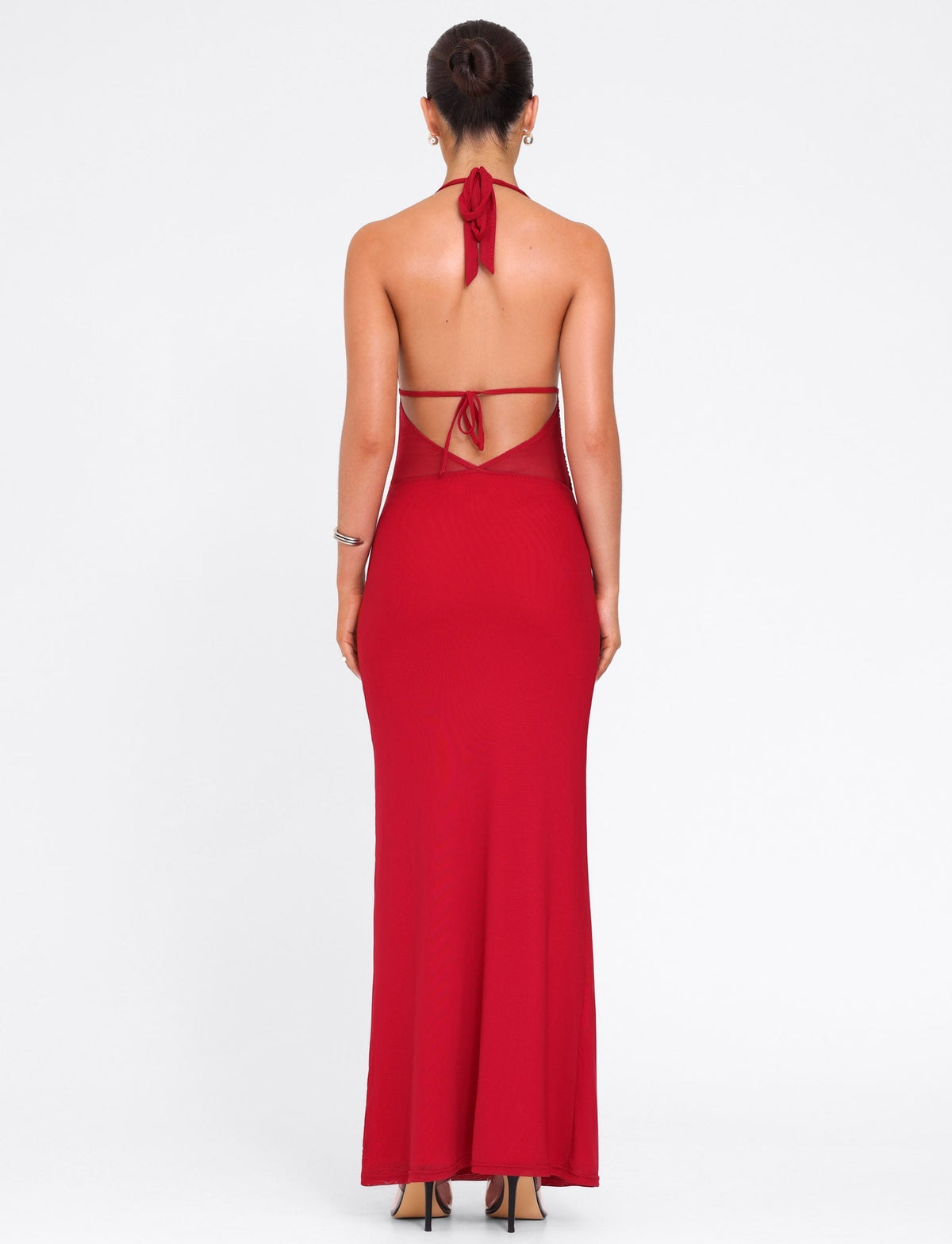 A woman stands facing away, wearing the TIGERMIST BRESILY MAXI DRESS in red, a stretch halter style with thin straps tied at the neck and lower back, paired with black heels; her hair is styled in a neat bun.