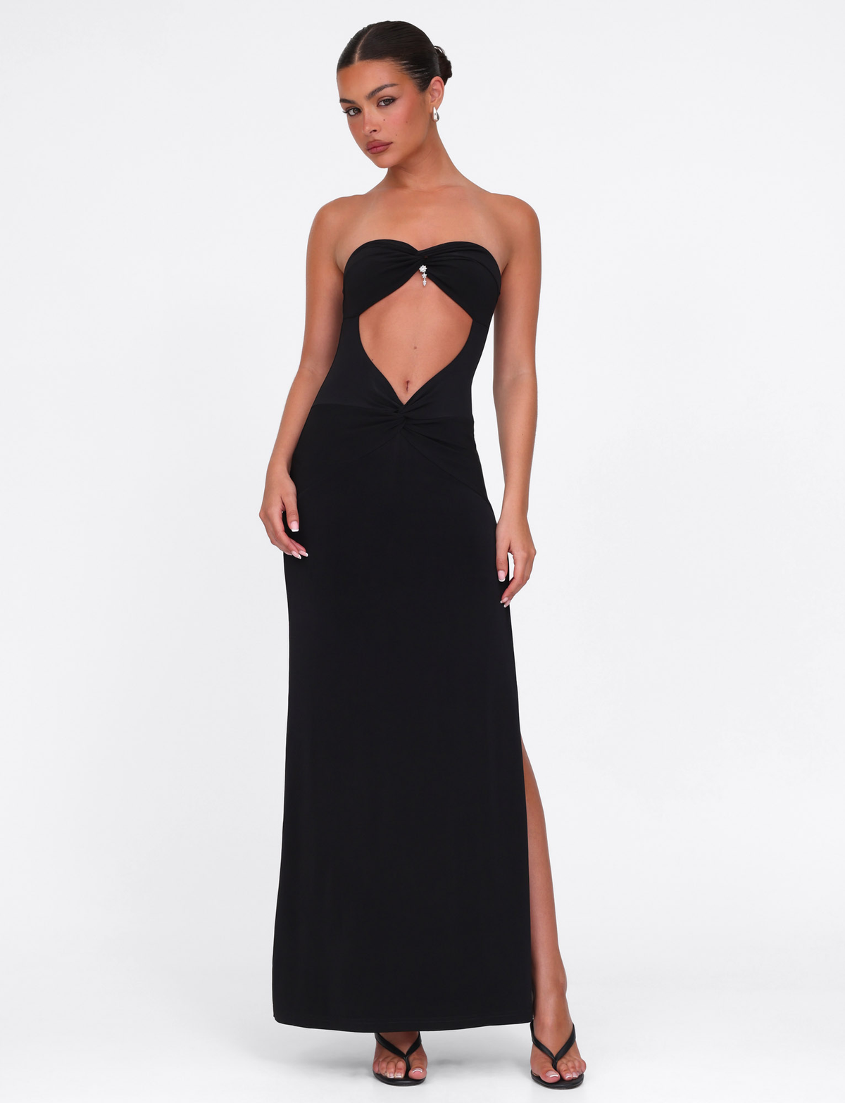A woman models the TIGERMIST GRAZIA DRESS - BLACK, featuring high stretch fabric, a bandeau neckline, front cutout, and high slit. She pairs it with black high-heeled sandals against a plain white background.