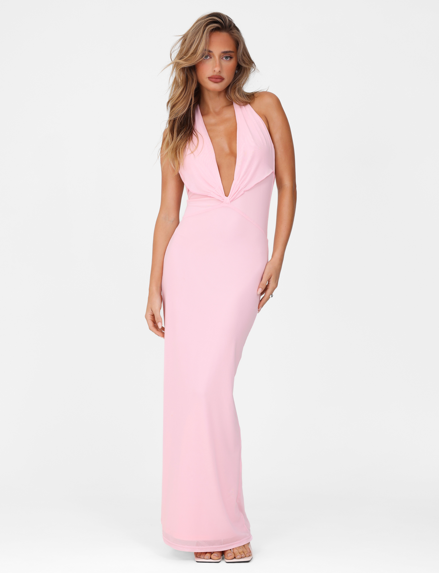 A woman wearing the TIGERMIST CRESSIDA MAXI DRESS in baby pink stands against a plain white background. The dress features a halter neck, plunging neckline, and maxi length. She has loose, wavy hair and wears strappy heels.