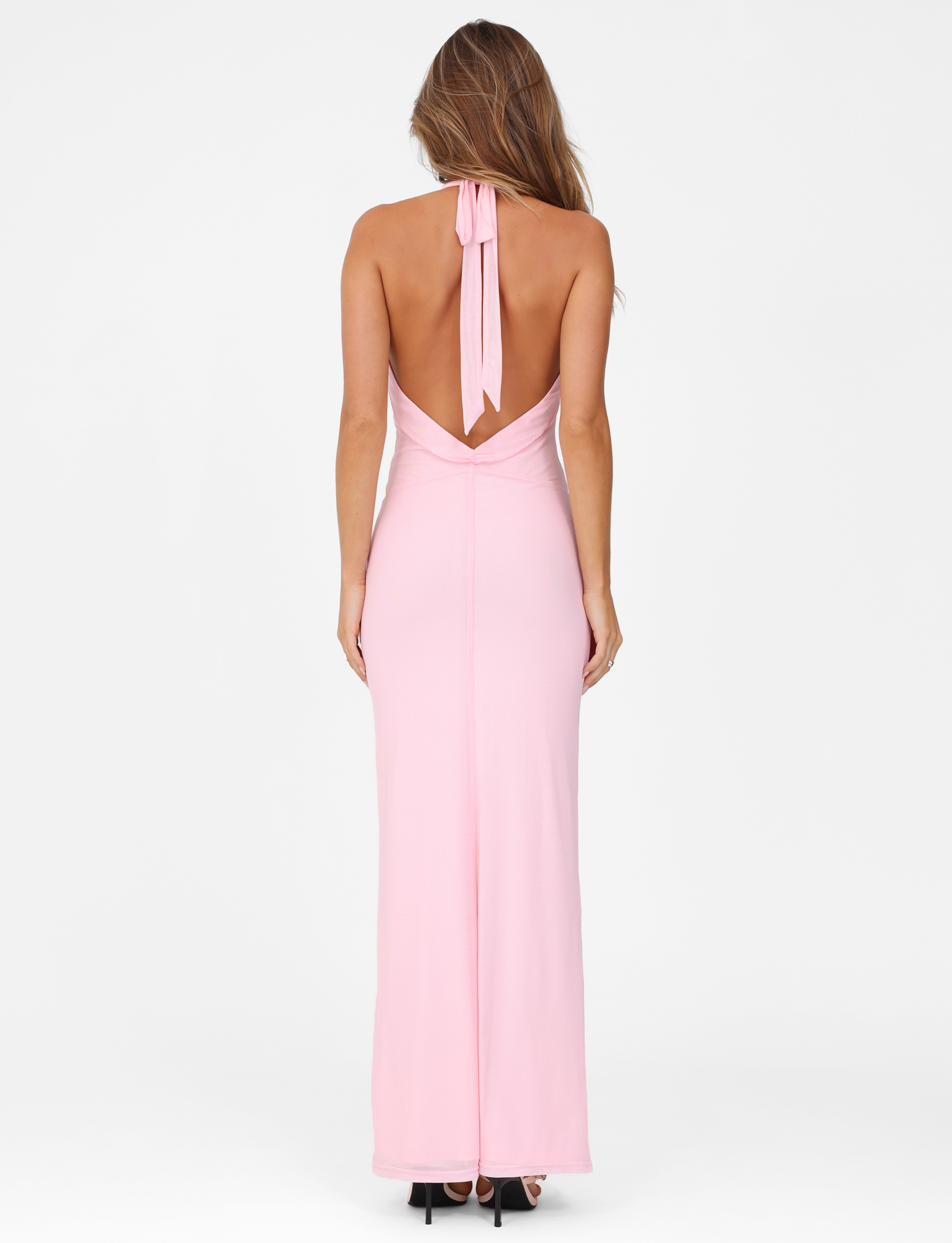 A woman with long, wavy hair models the TIGERMIST CRESSIDA MAXI DRESS in baby pink, a backless halter neck maxi, standing with her back to the camera against a plain white background.