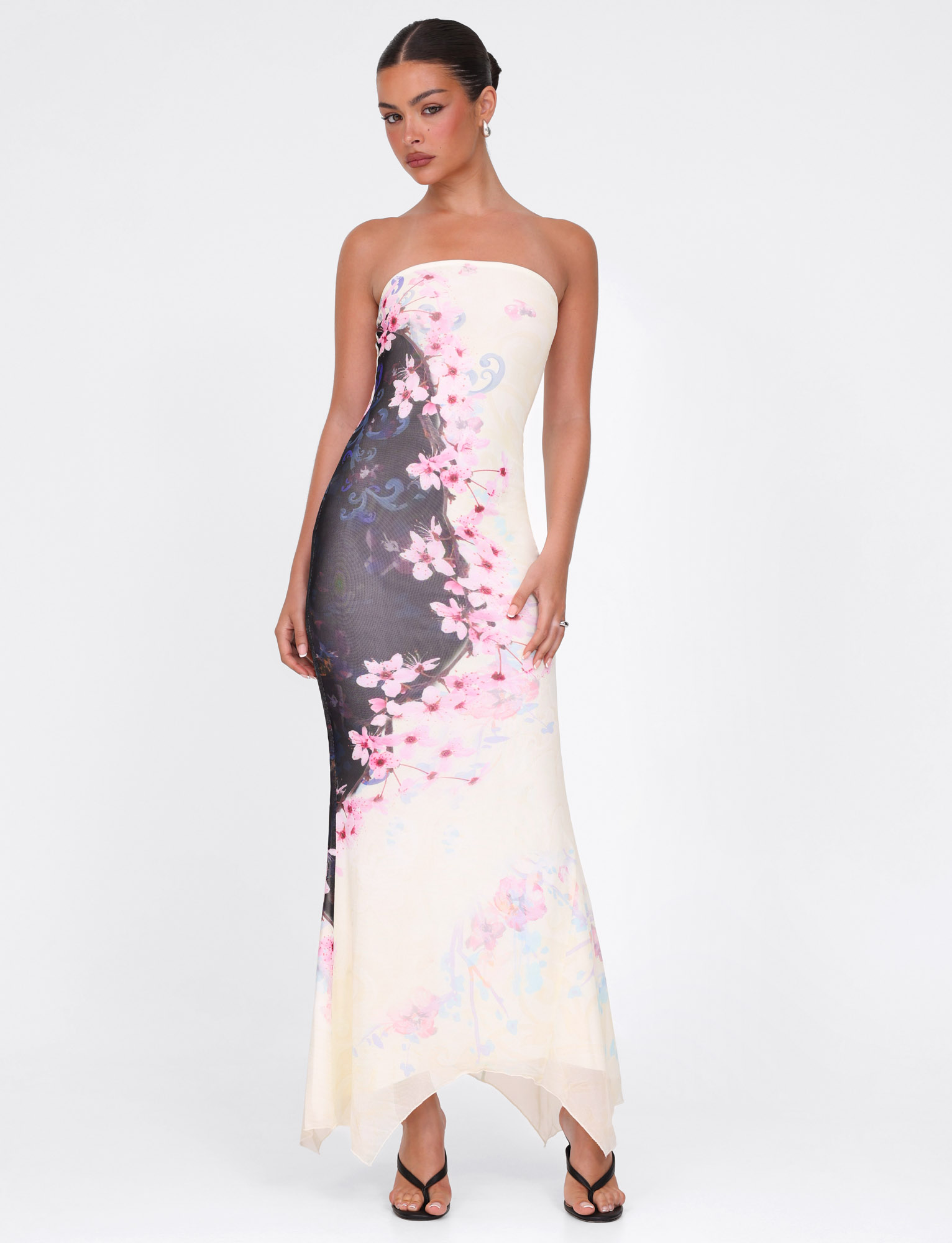 A woman stands against a white background wearing the TIGERMIST LILLIAN MAXI DRESS in multi floral cherry blossom print. Her hair is in a neat updo, and she pairs the strapless dress with black strappy heels.