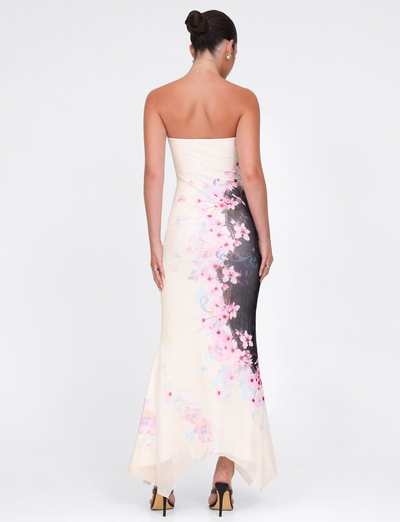A woman with her hair in a bun models the TIGERMIST LILLIAN MAXI DRESS in cherry blossom floral print (pink/black), standing against a white background and facing away from the camera.