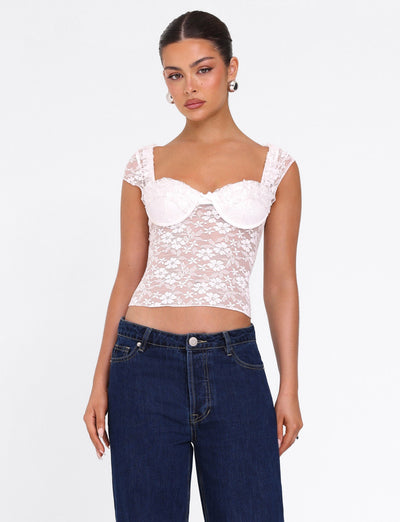 A woman stands against a white background wearing the TIGERMIST NYRA TOP in white, featuring lace fabric and sheer shoulder straps, paired with high-waisted dark blue jeans. Her hair is pulled back and she accessorizes with pearl earrings.
