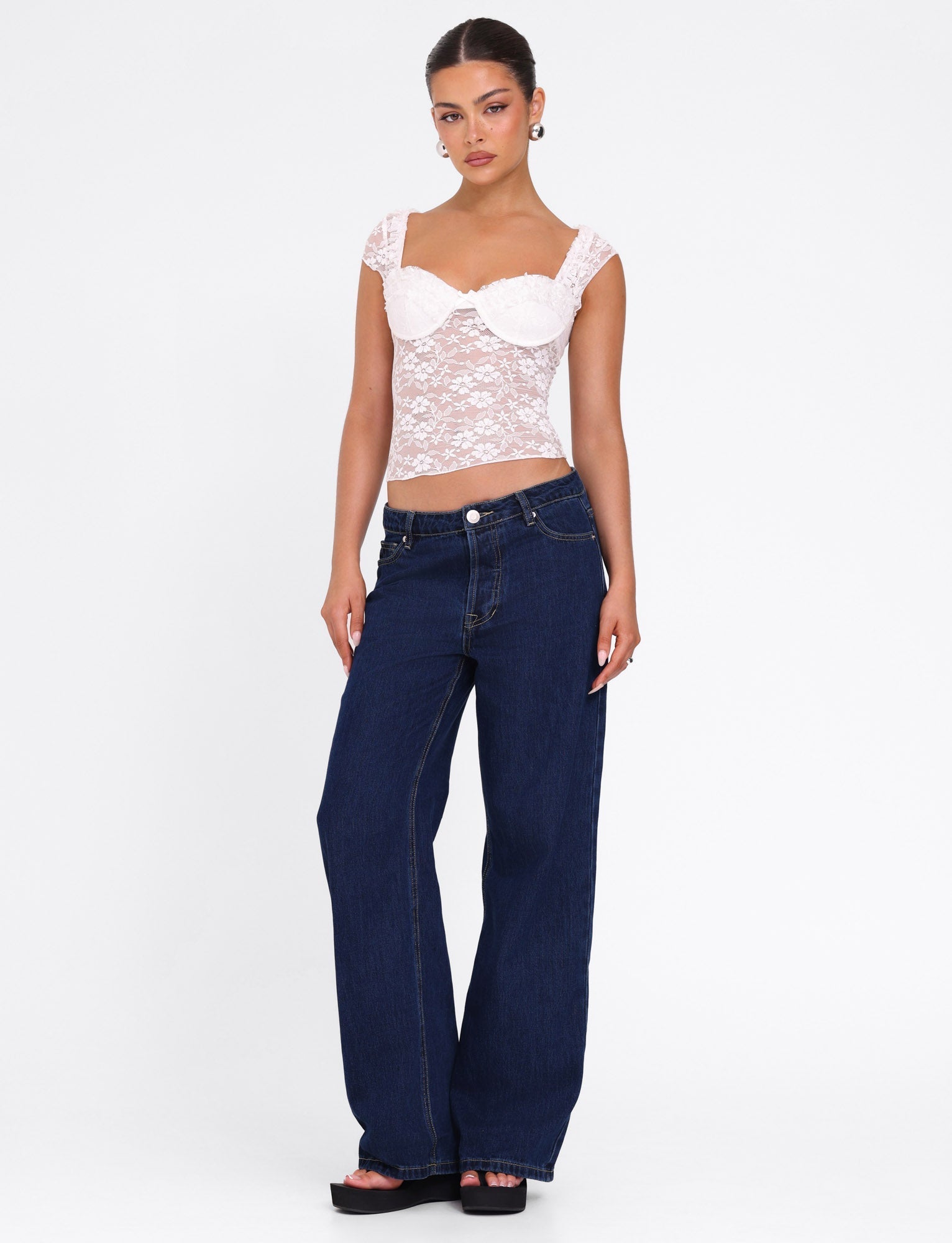 A woman stands against a white background wearing the TIGERMIST NYRA TOP in white, styled with wide-leg dark blue jeans, black platform sandals, hoop earrings, and her hair pulled back.