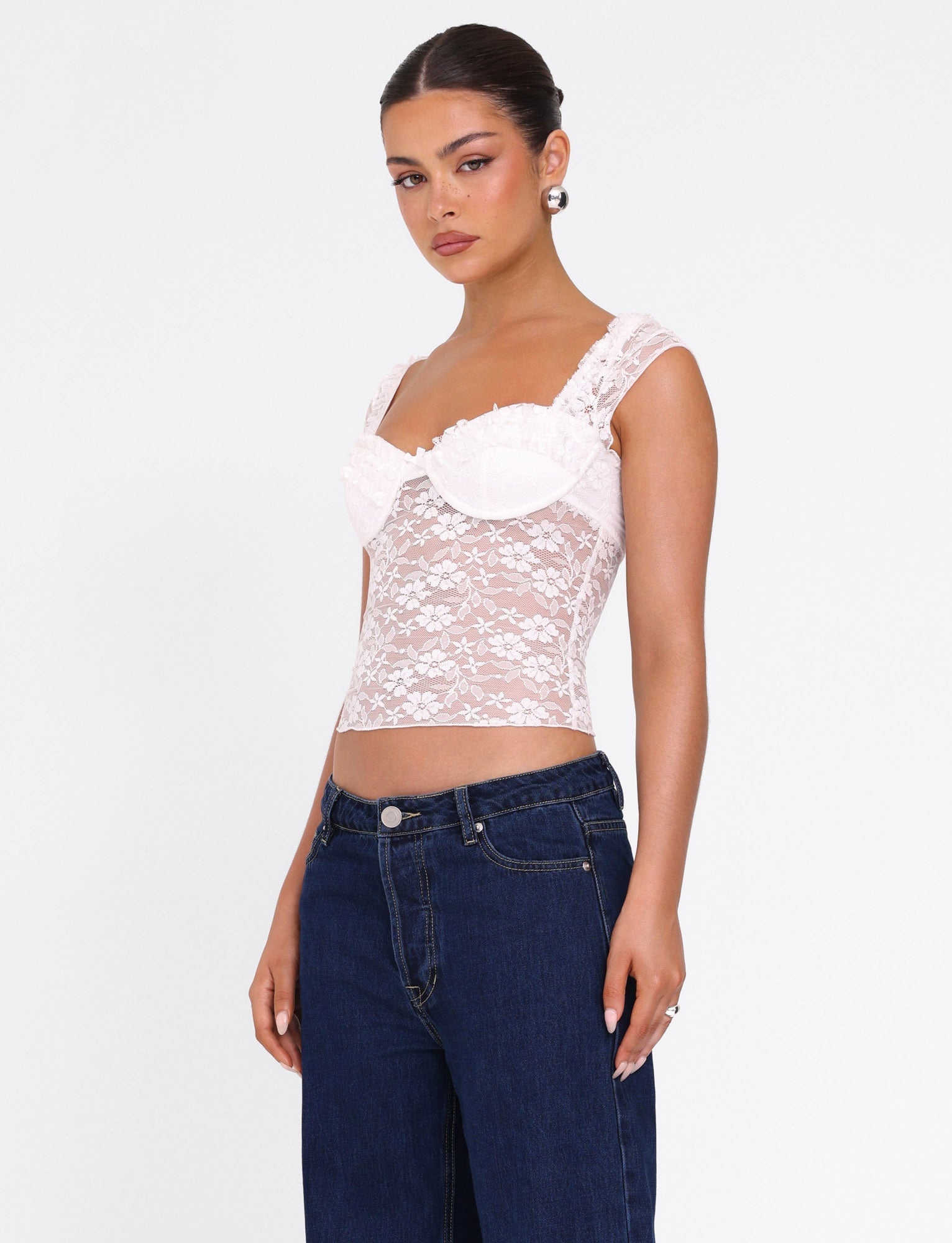 A woman models the TIGERMIST NYRA TOP - WHITE, a sheer white lace bustier with sheer straps, paired with dark blue jeans. She stands against a plain white background, her hair pulled back and expression neutral.