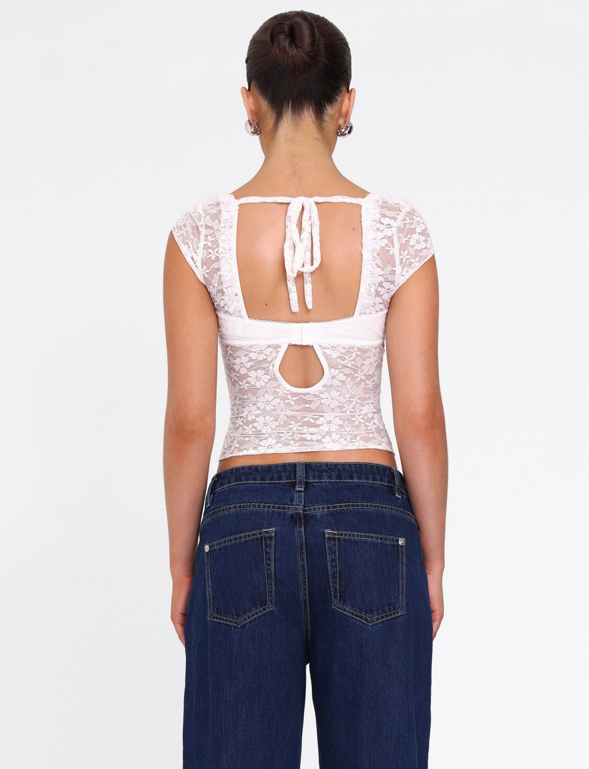 A woman with dark hair in a bun models the TIGERMIST NYRA TOP - WHITE, a short-sleeve lace bustier with open back and tie detail, paired with dark blue jeans. She faces away from the camera against a white background.