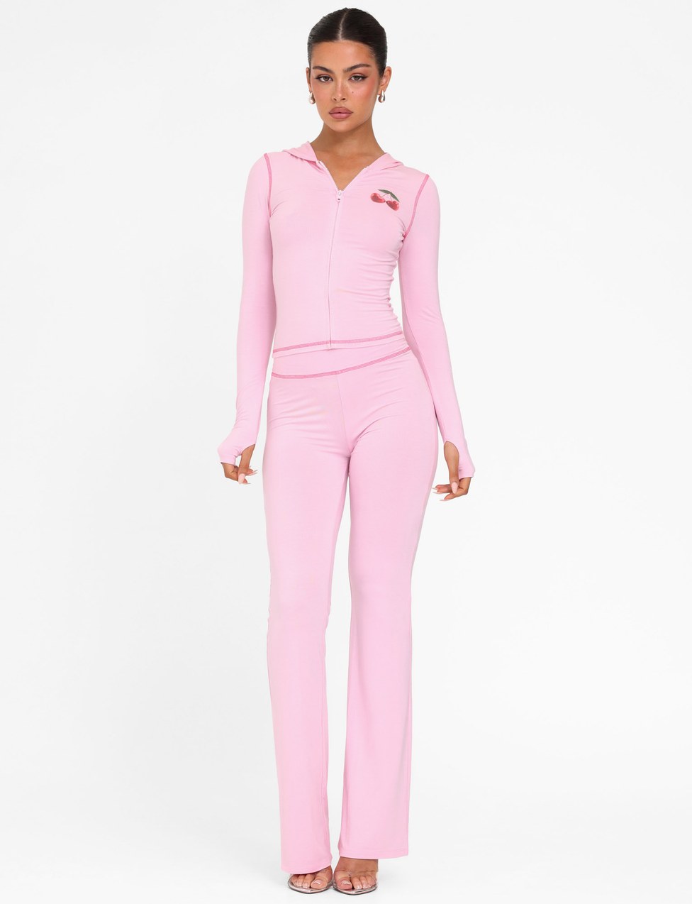 A woman faces forward in the TIGERMIST BLAYZE HOODIE - PINK (Soft Pink) and matching high-waisted, flared pants, with her hair tied back, hoop earrings, and open-toe heels against a plain white background.