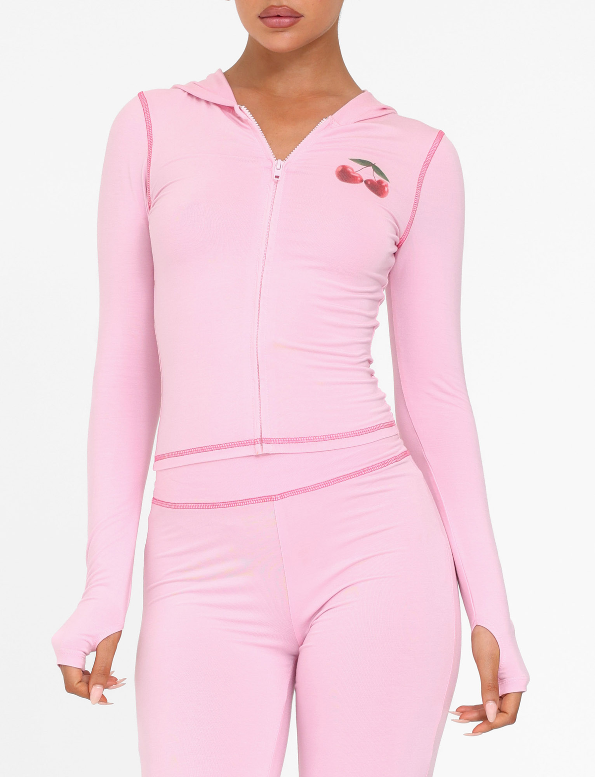 A woman wears the TIGERMIST BLAYZE HOODIE - PINK in soft pink, a fitted zip-up with thumb holes, long sleeves, and a small cherry graphic on the chest. She pairs it with matching pink leggings against a white background.