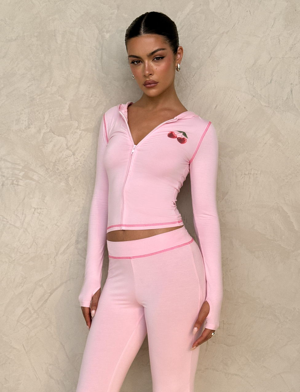 A woman in the TIGERMIST BLAYZE HOODIE - PINK stands against a textured beige wall, wearing soft pink zip-up hoodie with cherry chest graphic and matching high-waisted leggings, her hair slicked back and expression neutral.