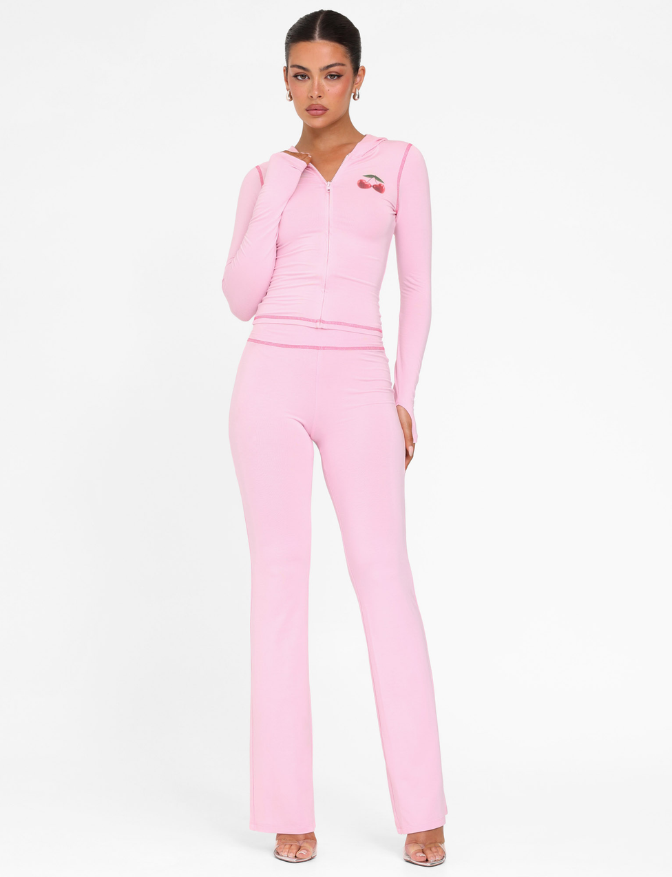 A woman poses against a white background in the TIGERMIST BLAYZE PANT - PINK : SOFT PINK, paired with a matching zip-up hoodie and heels, her hair pulled back.