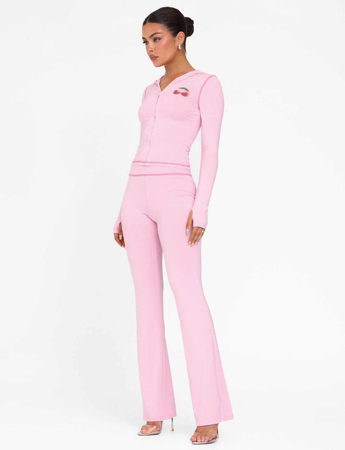 A woman stands against a white background wearing the TIGERMIST BLAYZE PANT in soft pink with a matching fitted zip-up top, her hair styled in a neat bun, and clear high-heeled sandals.
