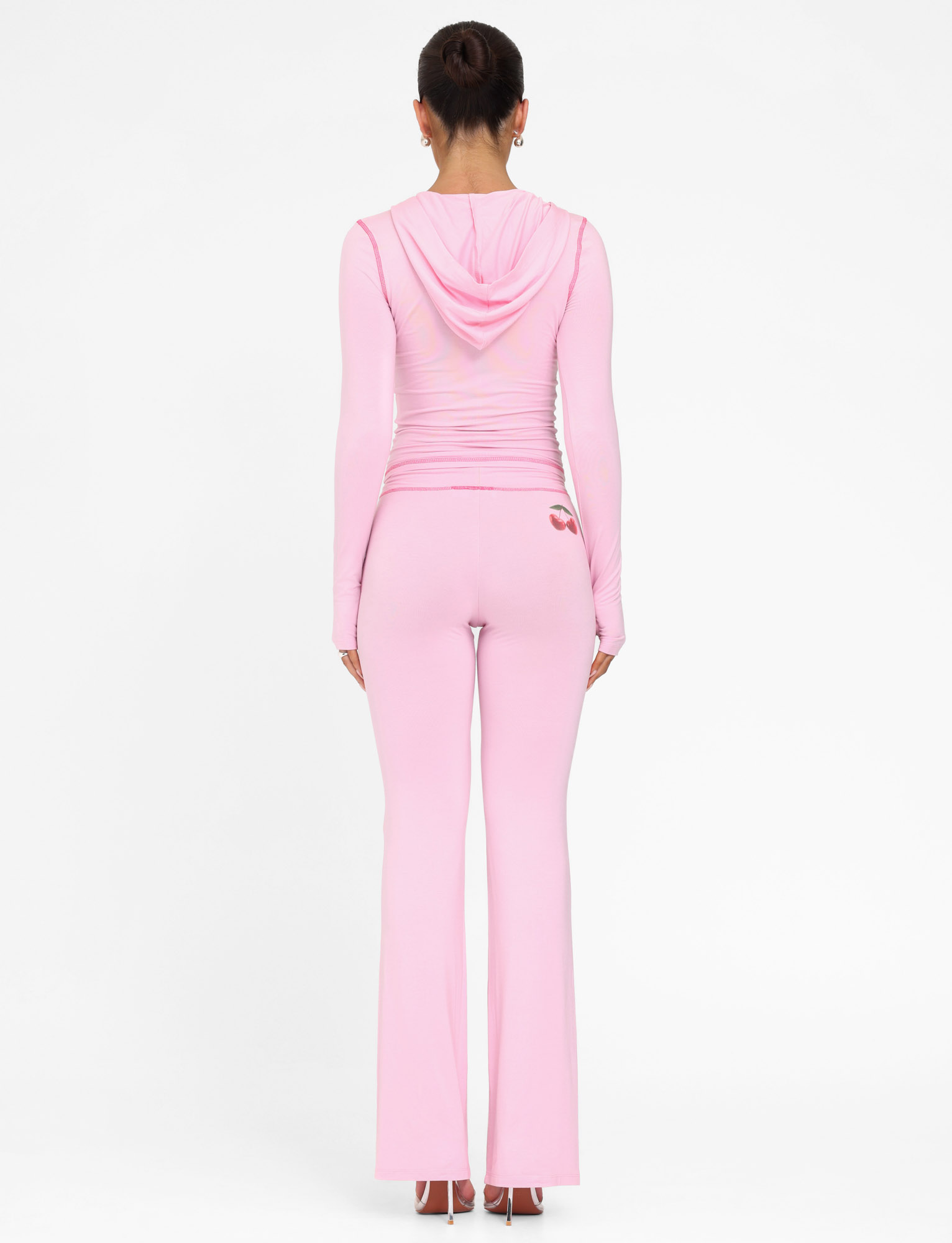 A woman stands facing away, wearing TIGERMIST's BLAYZE PANT in soft pink with a matching fitted light pink hooded top, her hair in a neat bun and arms relaxed by her sides, set against a plain white background.